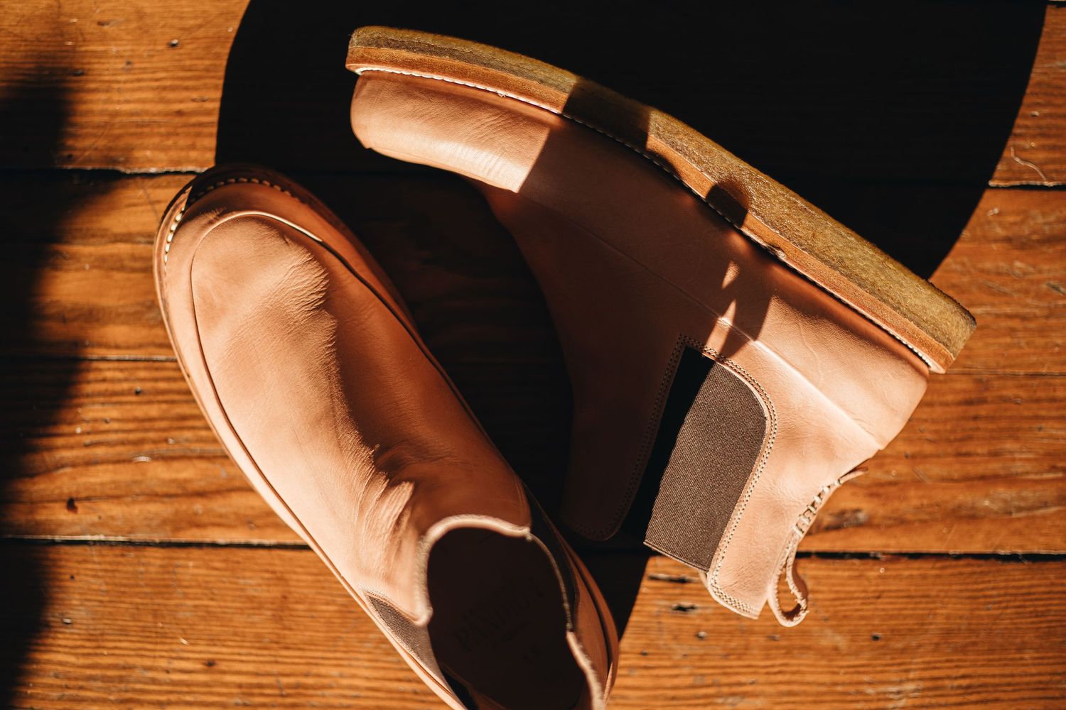 Tan leather Chelsea boots with elastic side panels resting on a rustic wooden floor in warm sunlight.