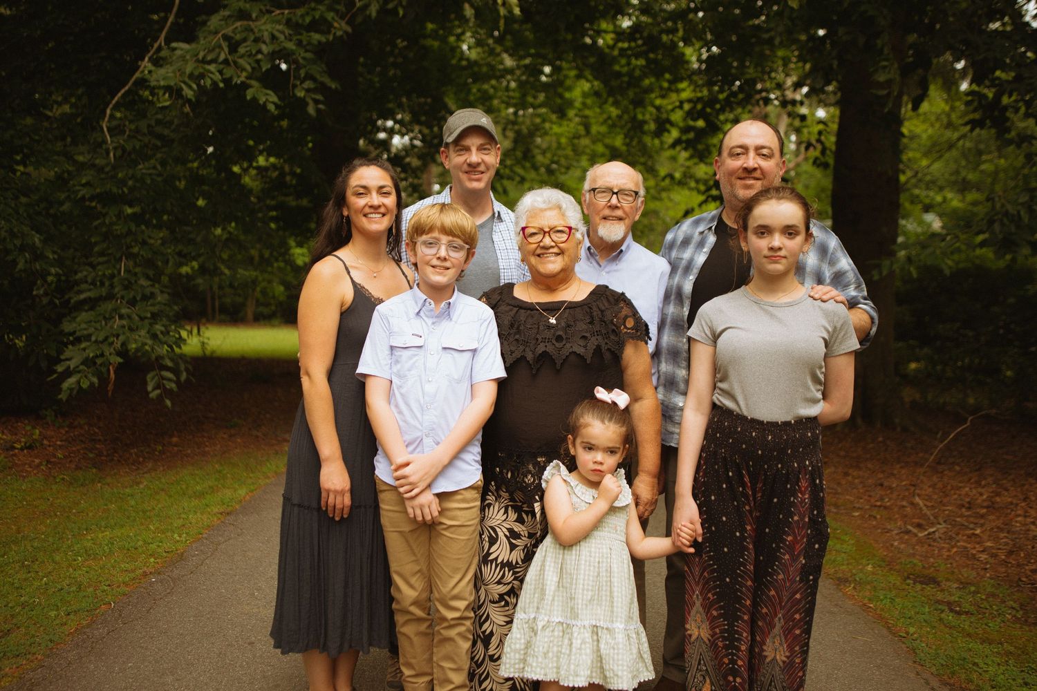Multi-generational family portrait outdoors on a tree-lined path, featuring grandparents, parents, and children smiling together.