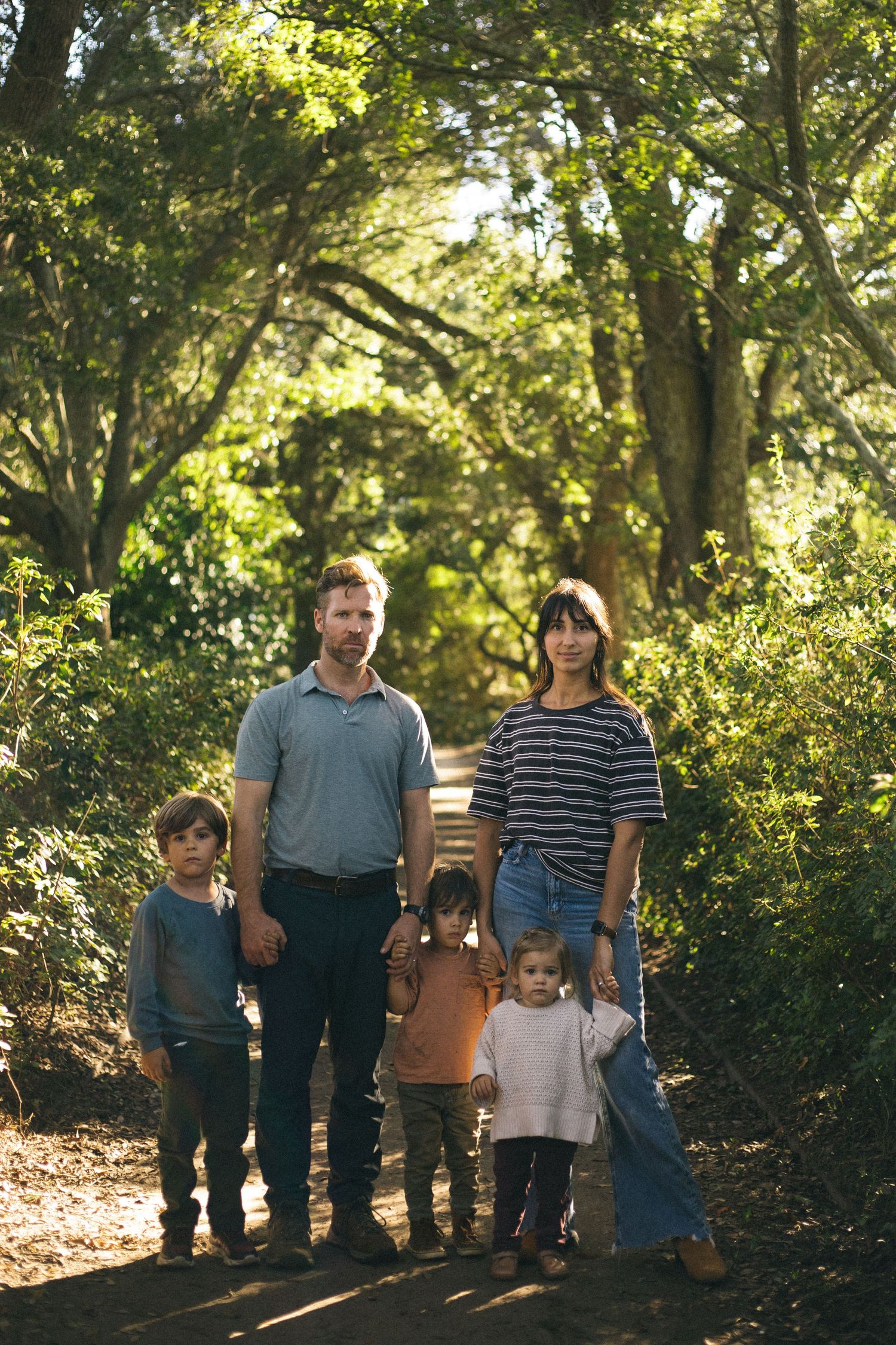 Family of five posing on a sunlit forest trail surrounded by lush green trees.