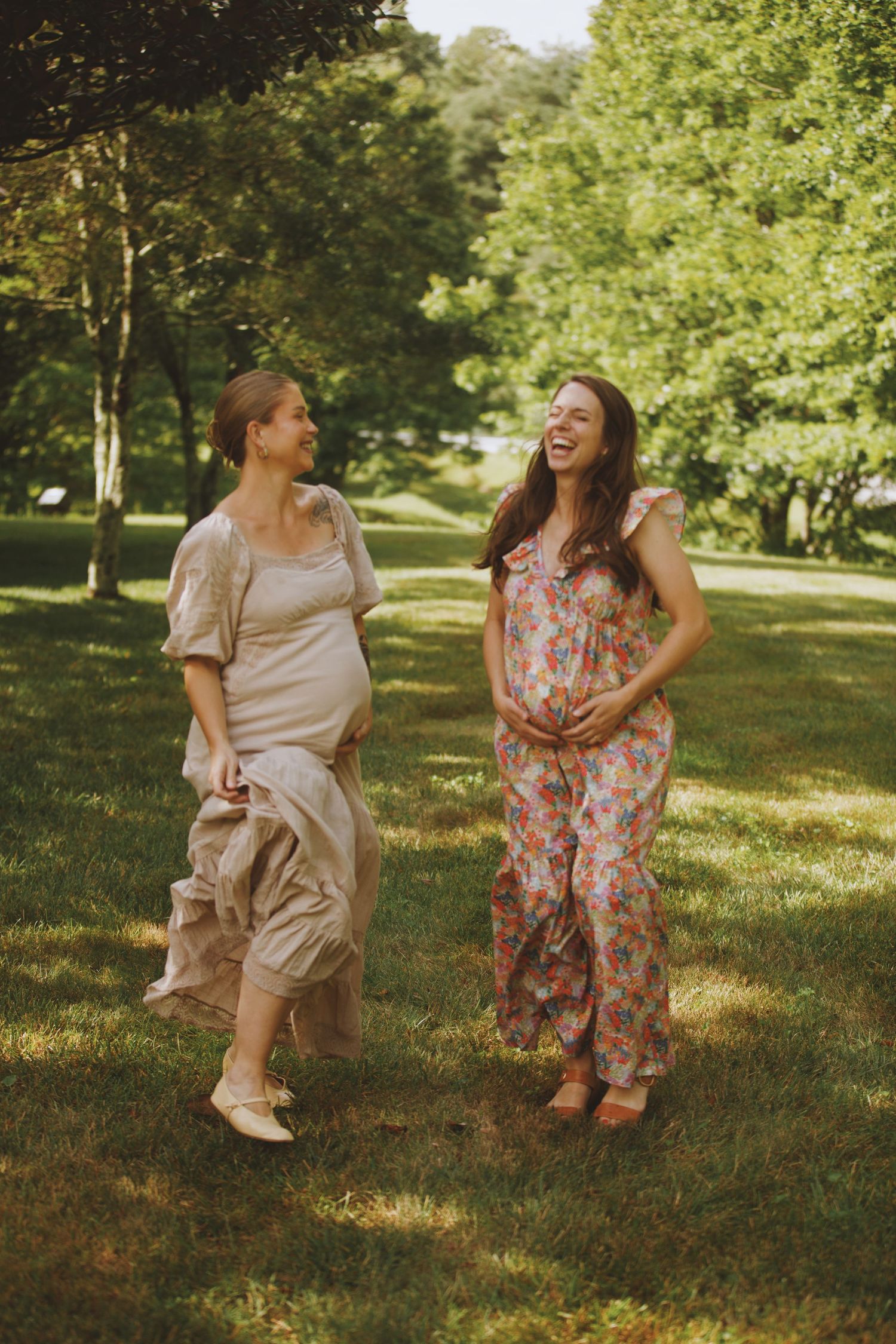 Two pregnant women laughing and walking together outdoors in a sunny green park, wearing summer dresses.