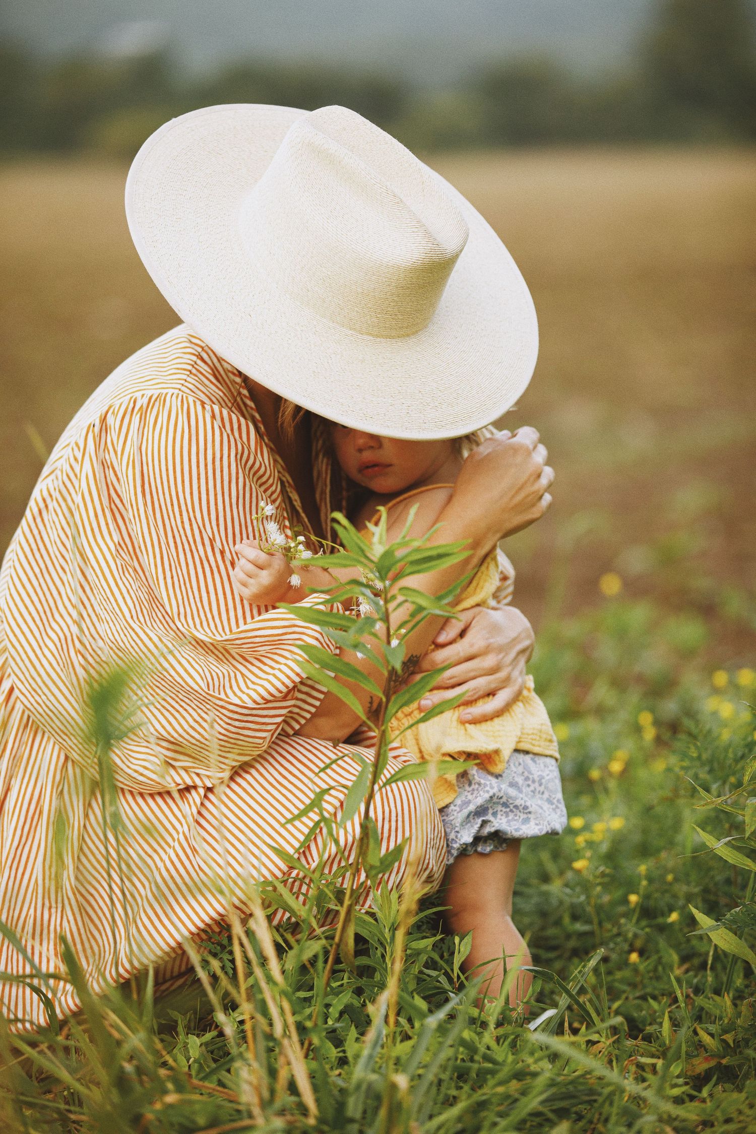 Woman in striped dress and white hat crouching in a field, tenderly embracing a small child among green grass.