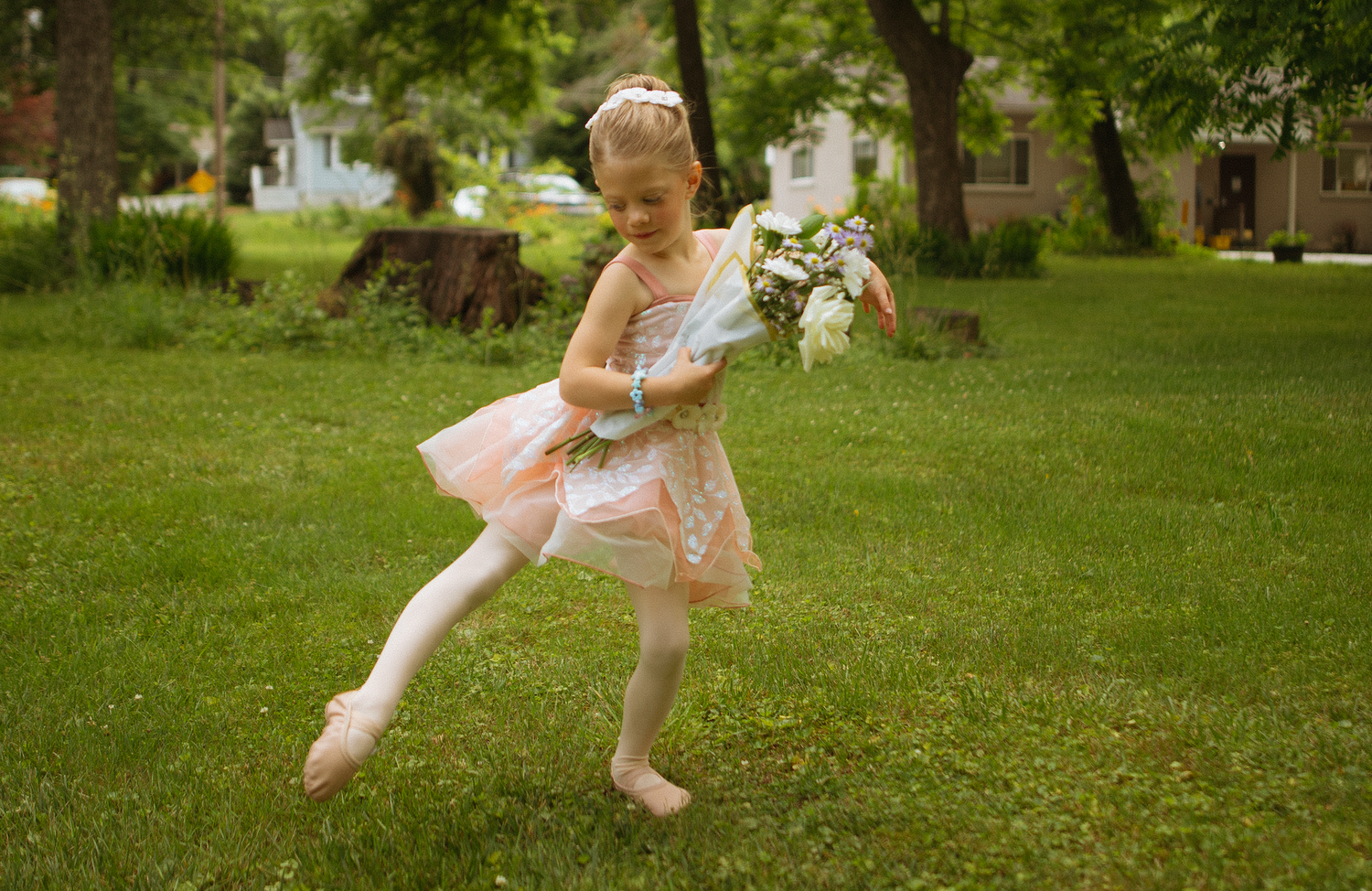 Young girl in pink ballerina dress and tights holds white flowers while dancing on green grass outdoors.