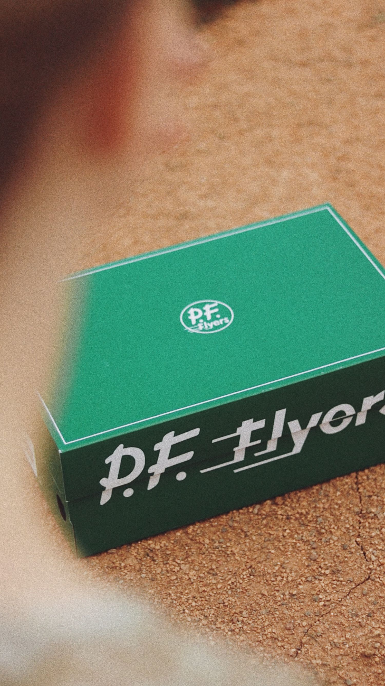 Green P.F. Flyers shoe box on a cork surface, photographed from above with shallow depth of field.