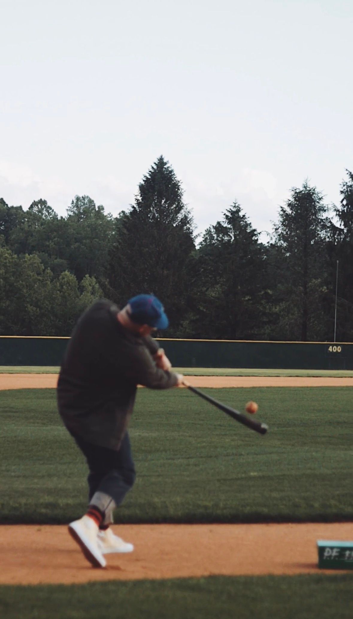 A baseball player in a blue cap swings a bat at a ball on a green field with trees in the background.