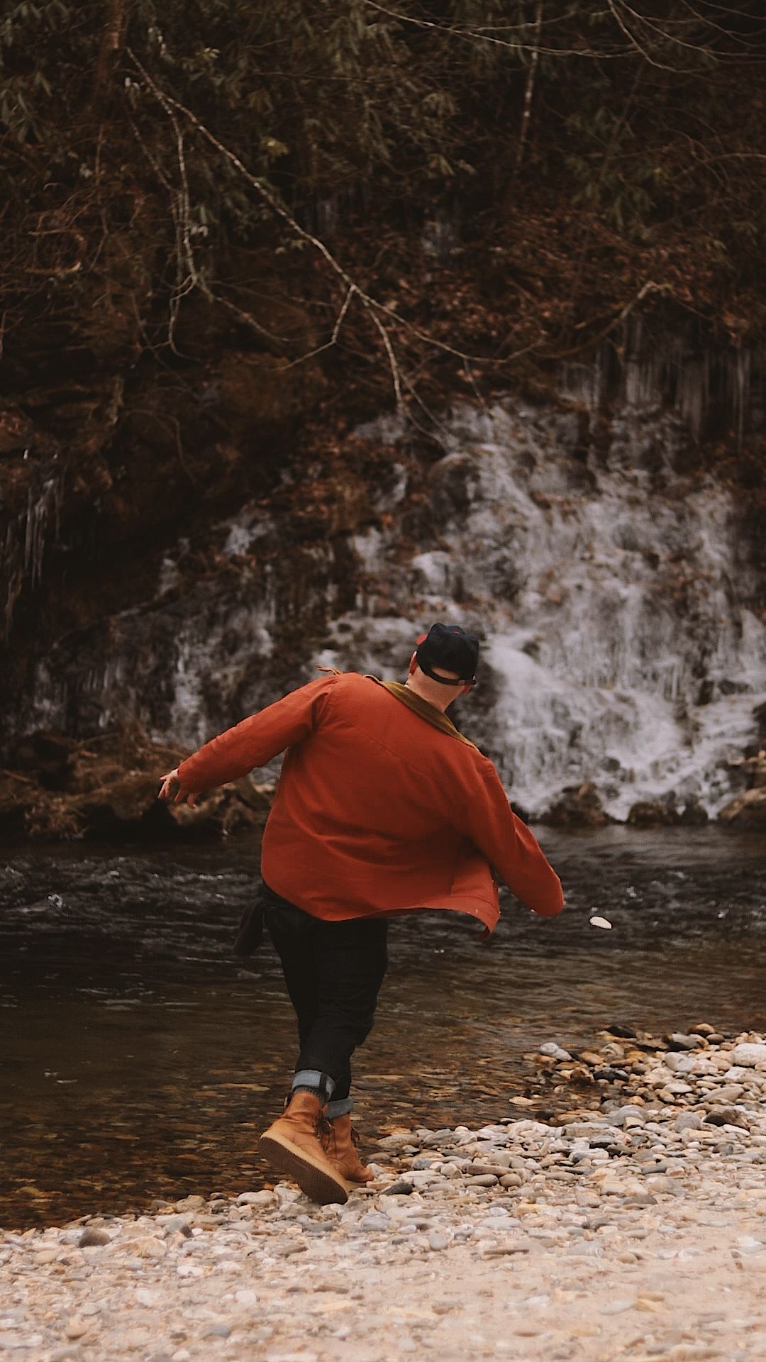 Person in red jacket and yellow boots skipping stones by a rocky stream with icy waterfall backdrop.