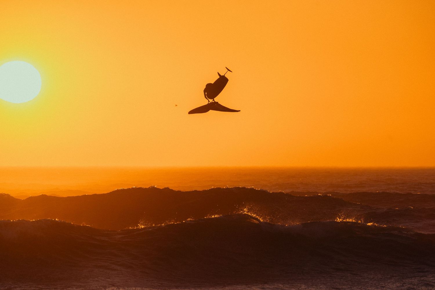 Silhouette of a pelican flying against vibrant orange sunset sky over mountain landscape.