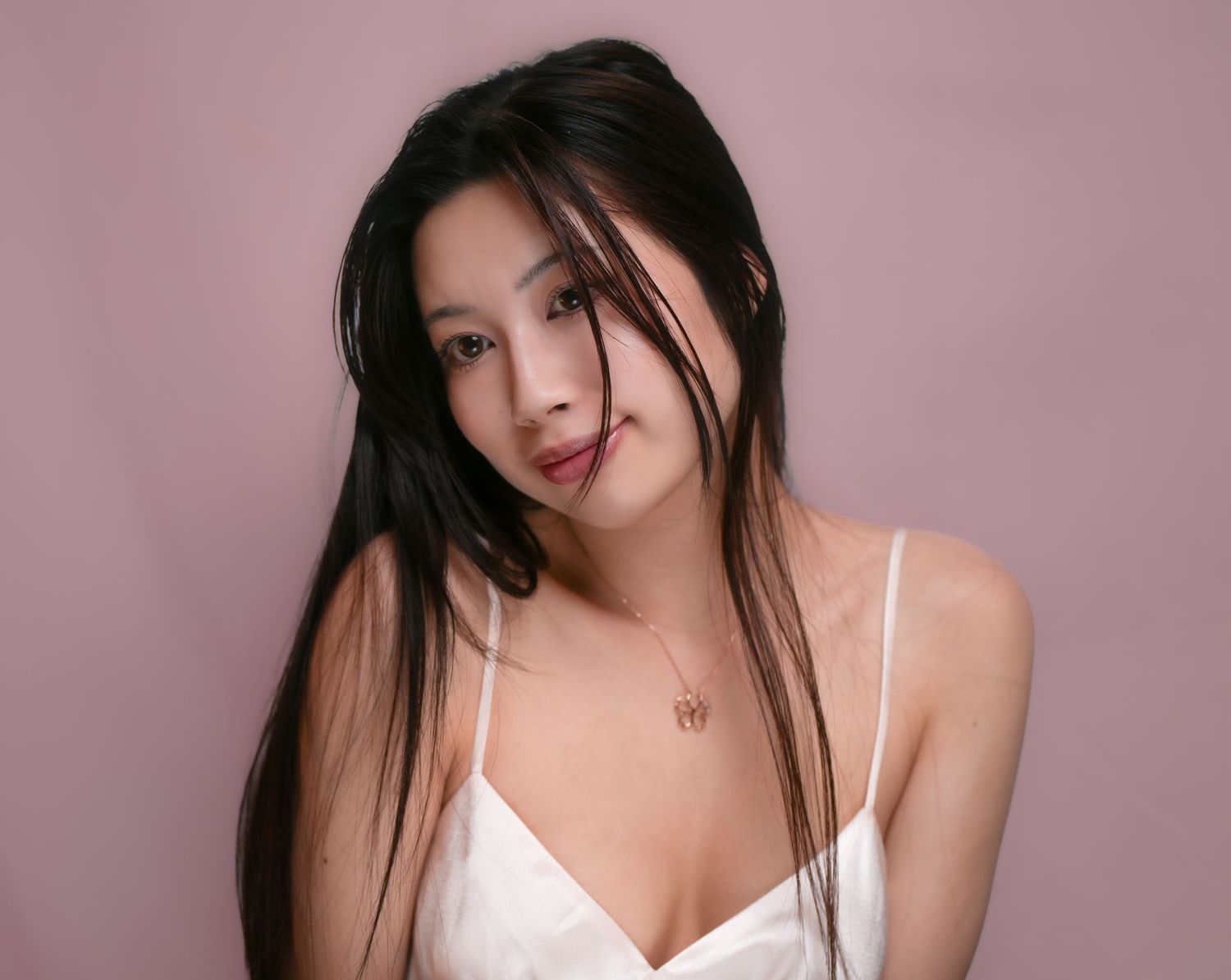 Woman with long dark hair wearing a white camisole poses against pink background in studio portrait.