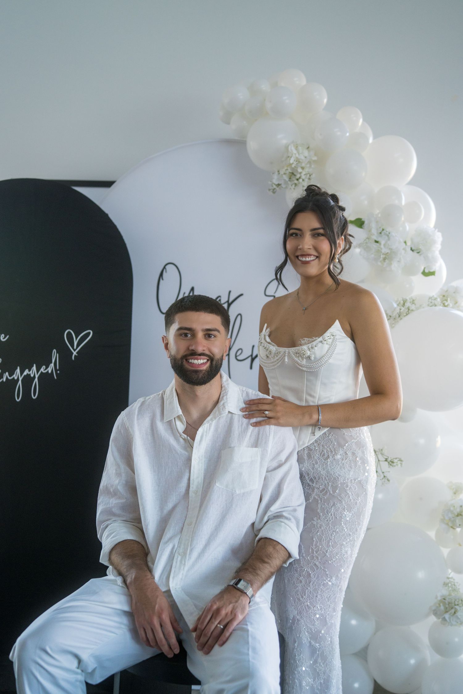 Modern wedding engagement photo shoot with white balloon arch and elegant white attire against dark backdrop.