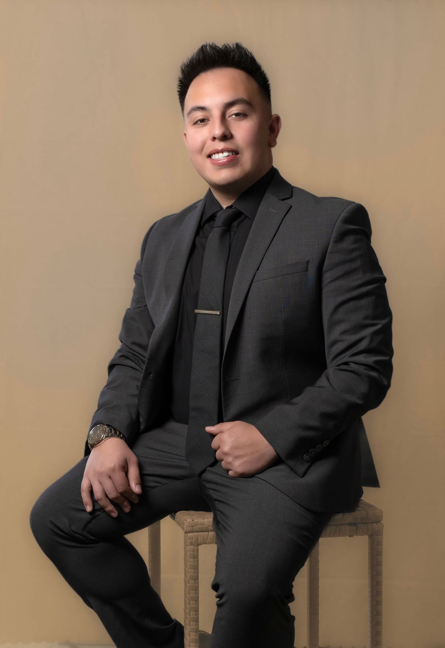 Professional studio portrait of a person in a sleek black suit and tie seated against a neutral backdrop.