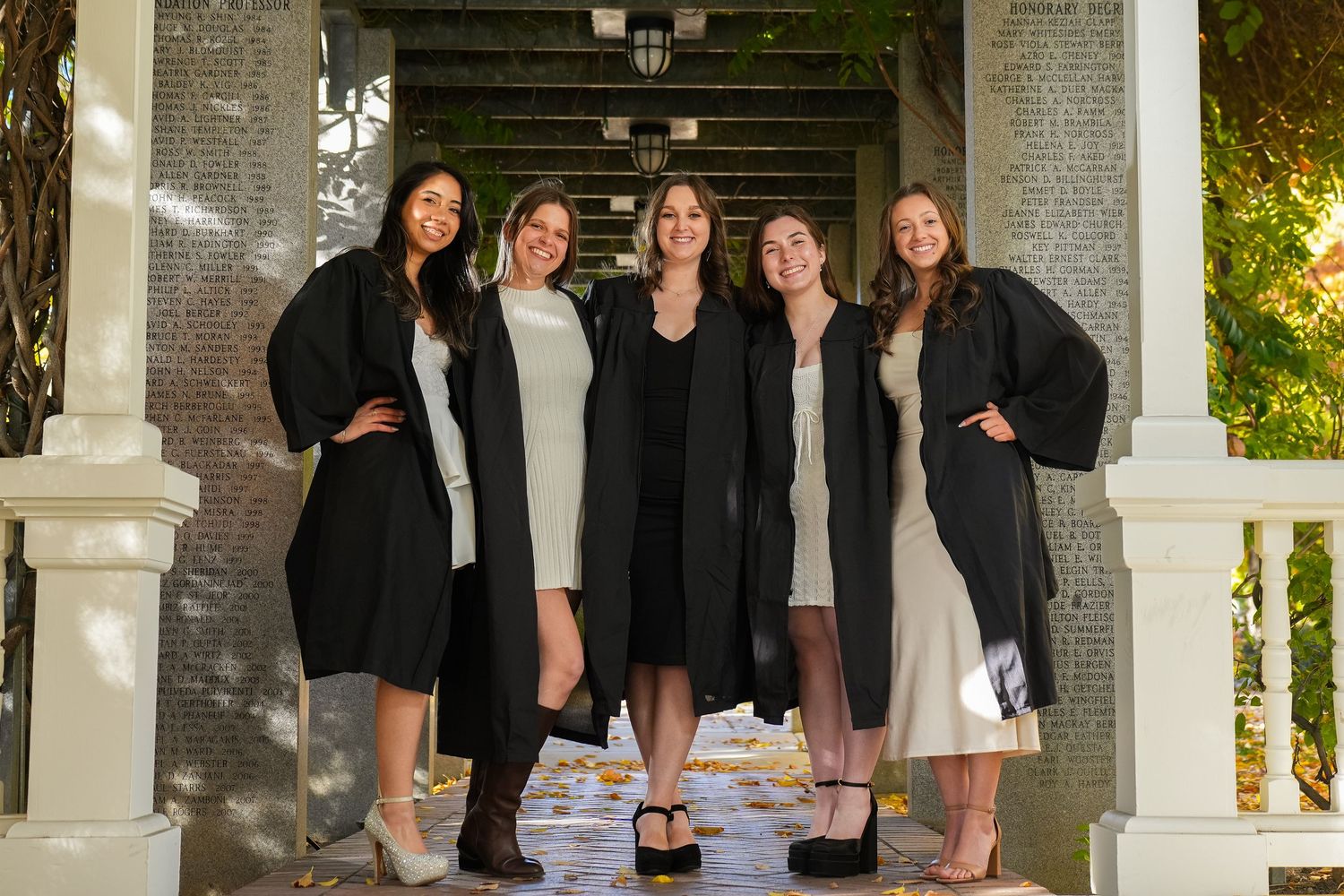 Group of graduates in black and white dresses pose together under columned walkway.