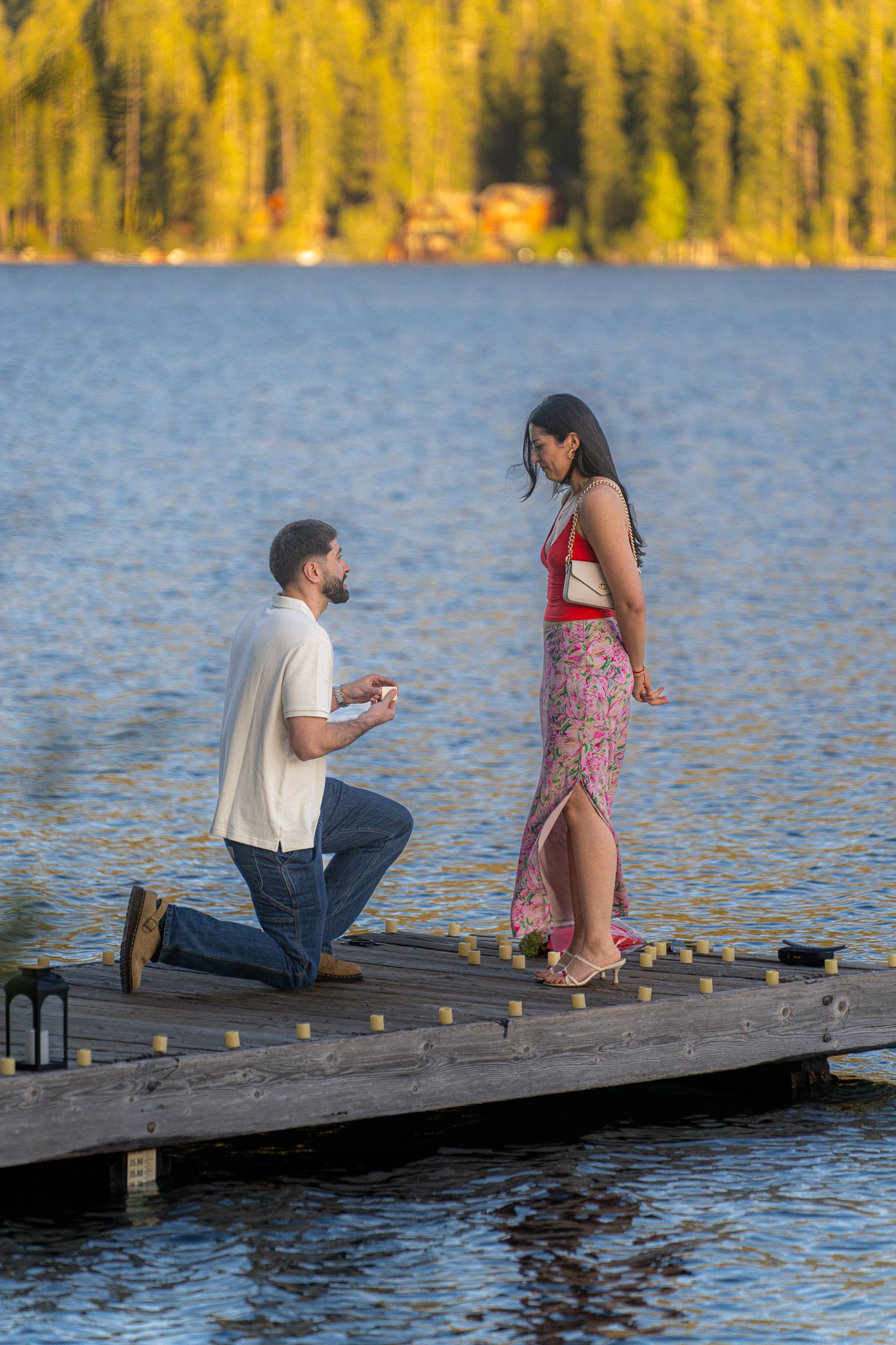 Two people share a romantic moment on a wooden dock by a lake with golden autumn trees in the background.