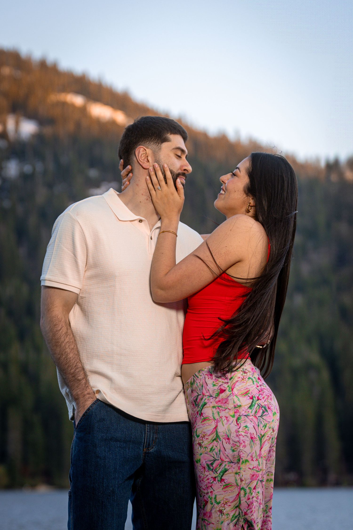 A romantic couple shares joyful laughter and tender moments while posing outdoors near a lake with mountains in the background.