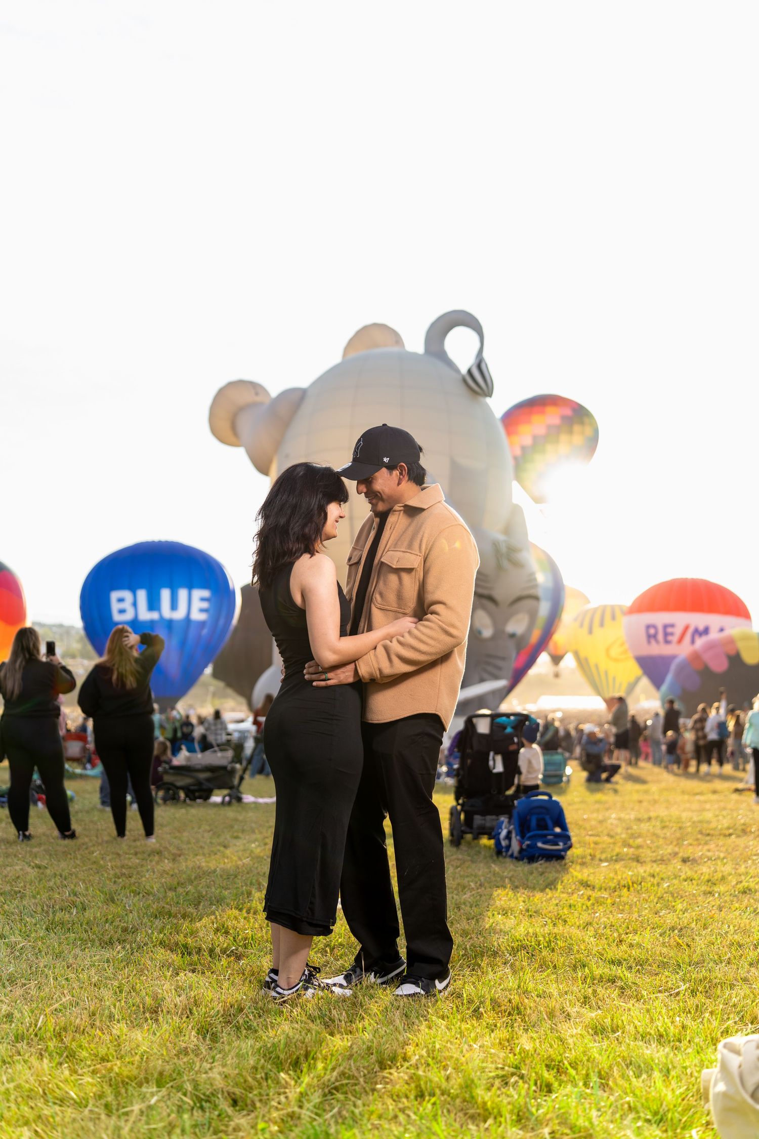 Hot air balloons inflate at dawn as couples gather on a grassy field during a balloon festival.
