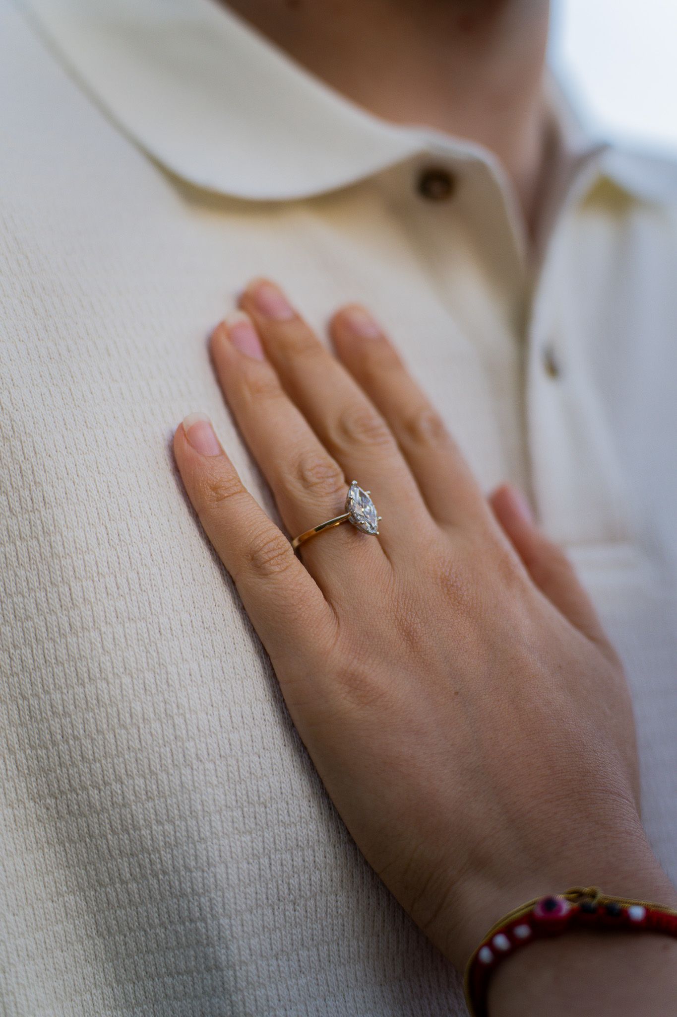 A delicate diamond engagement ring shown on a hand resting against a cream-colored collared shirt.