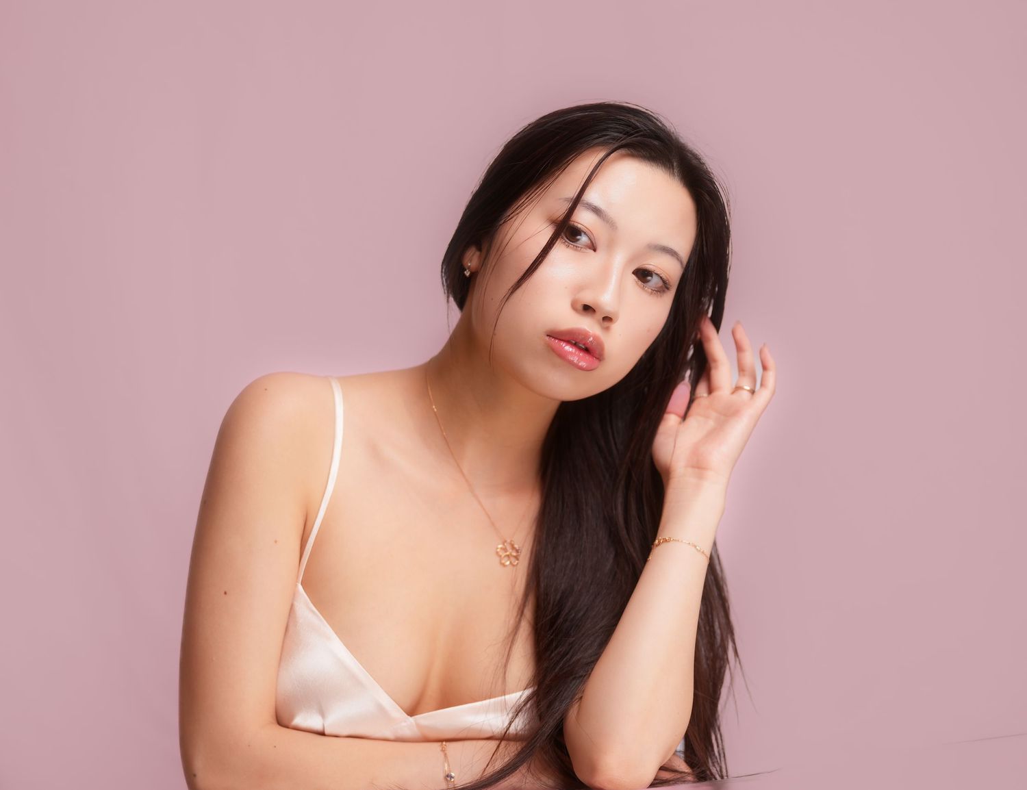 Asian woman with long dark hair in white top poses against pink background while touching her hair.