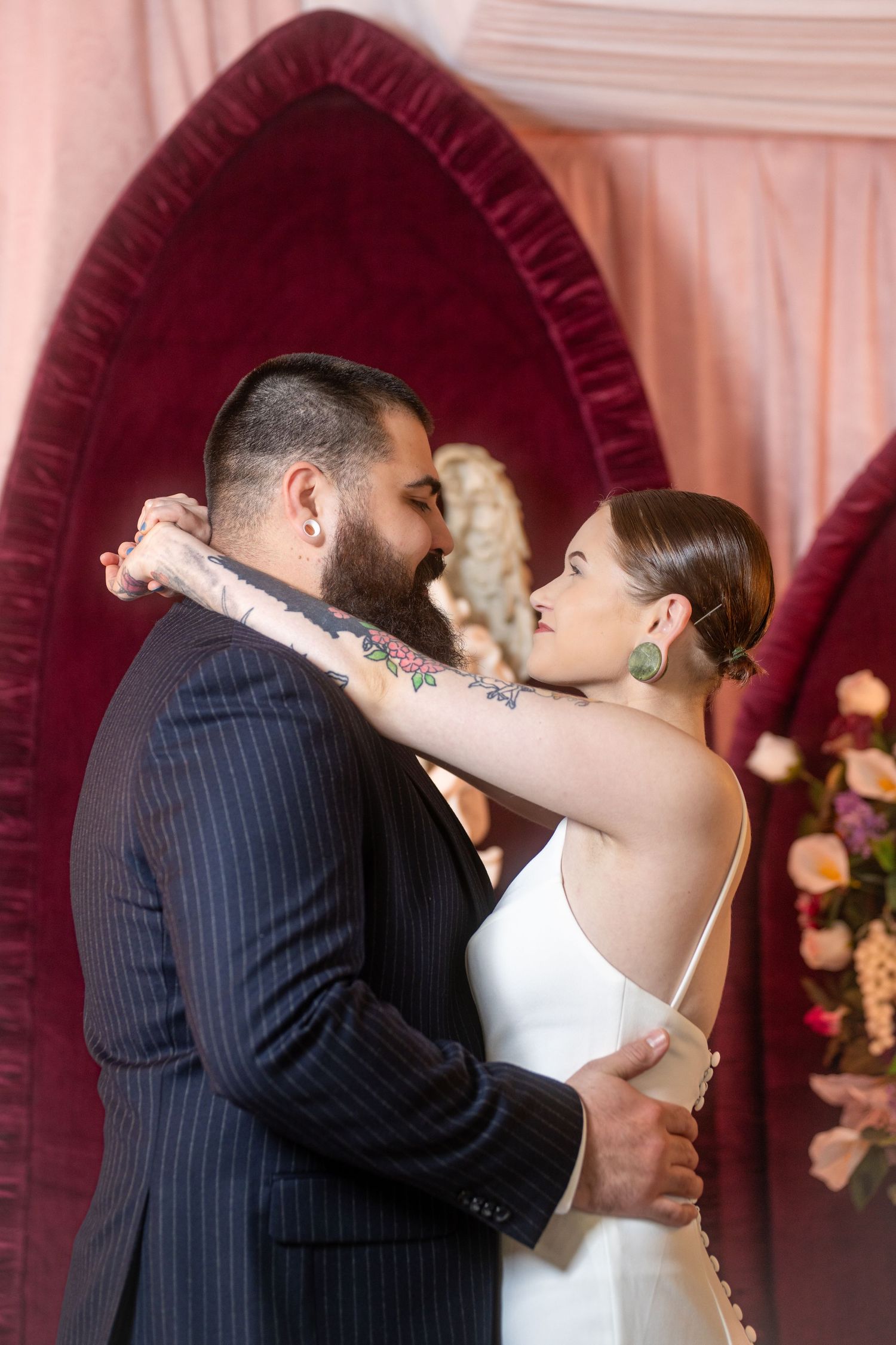 A couple shares an intimate moment during their wedding ceremony in front of a burgundy velvet arch with floral decor.