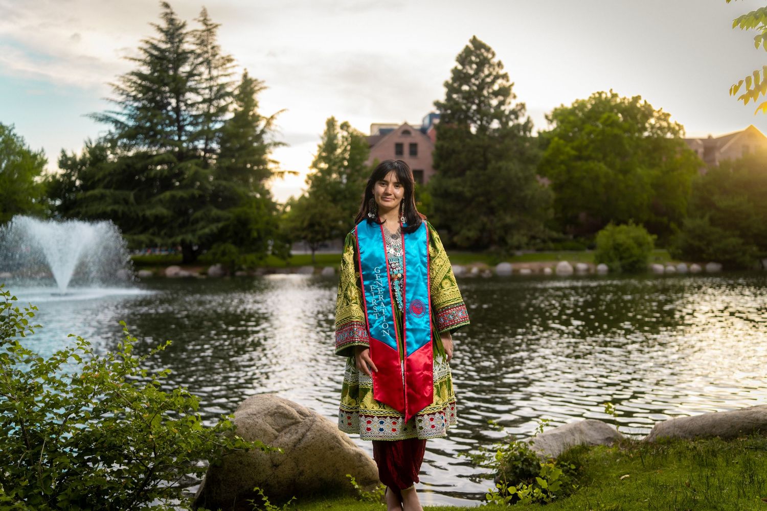 Someone stands near a scenic pond with a fountain at sunset, surrounded by evergreen trees and rocks.