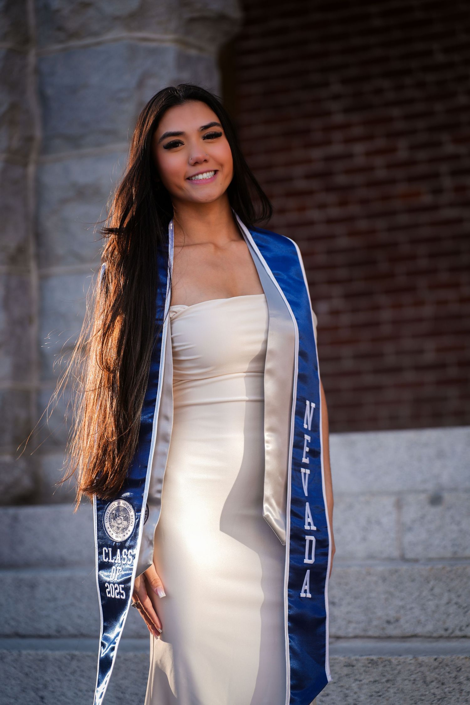 Graduate in white dress and blue Nevada stole poses against brick wall.