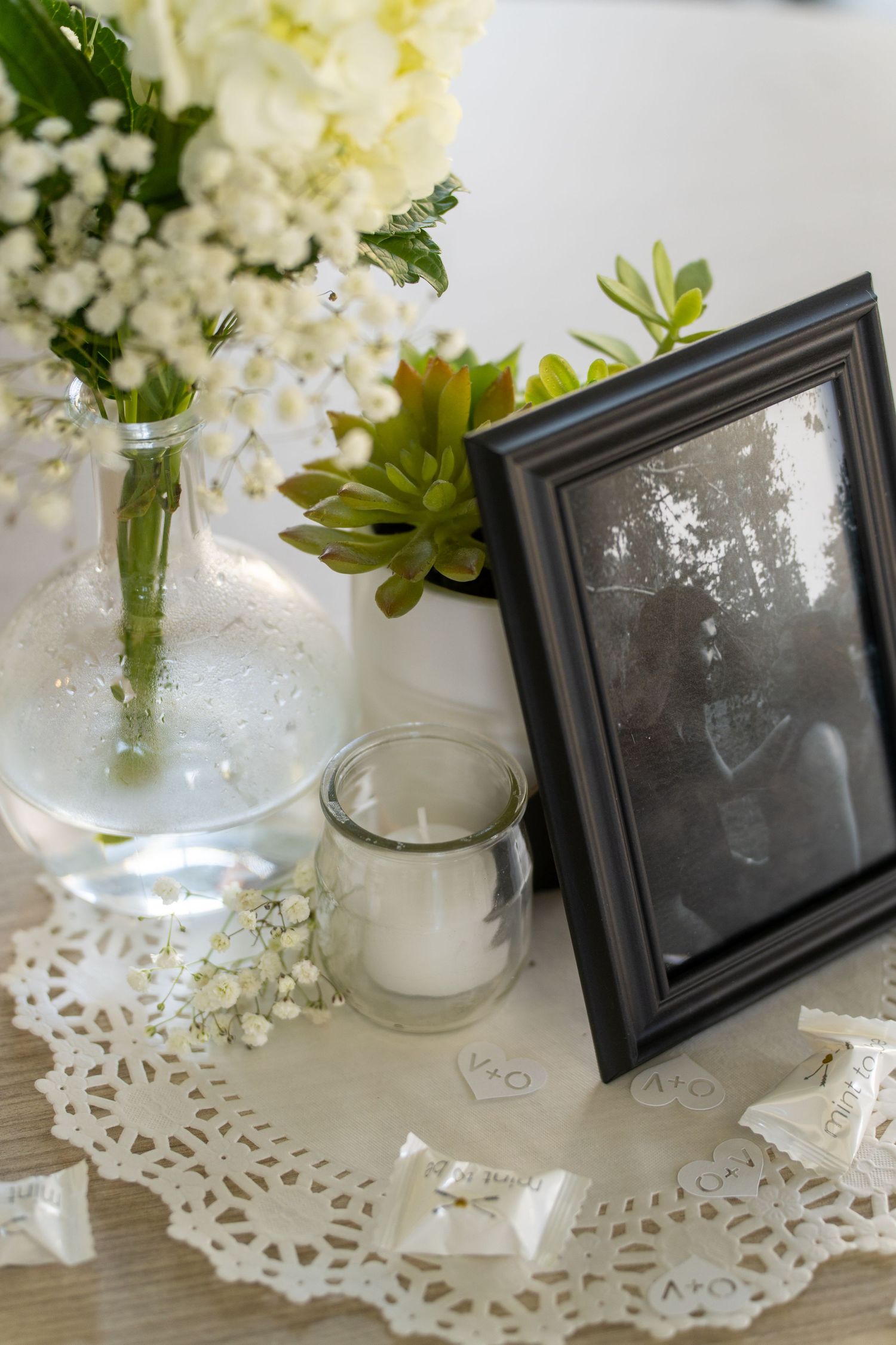 White flowers in a glass jar sit beside a black frame on a delicate lace doily against a light background.