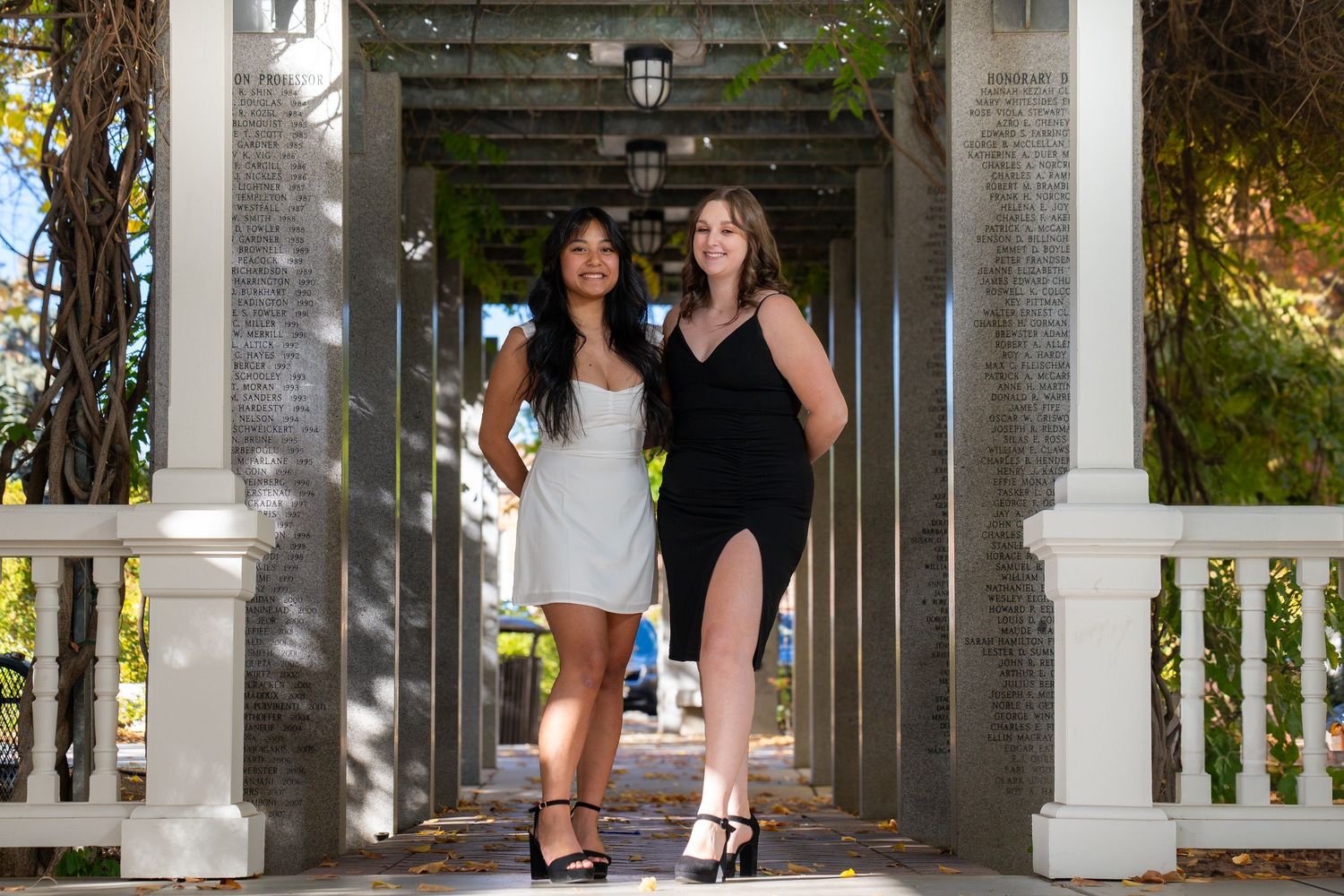 Two graduates posing in a white columned walkway wearing formal dresses and graduation stoles.