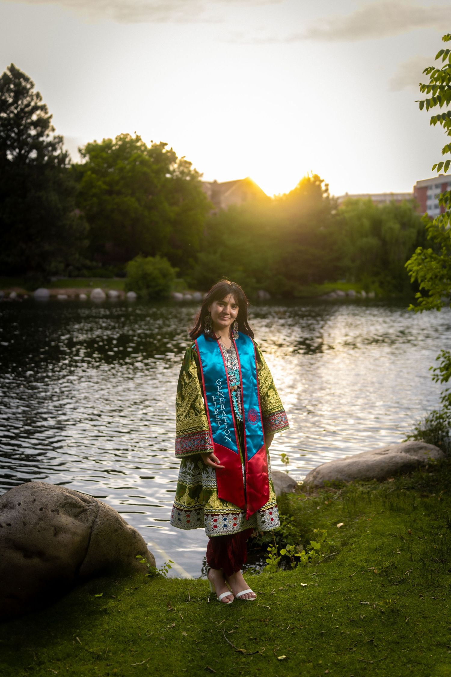 Person in colorful traditional dress stands by a serene lake at sunset with trees in the background.