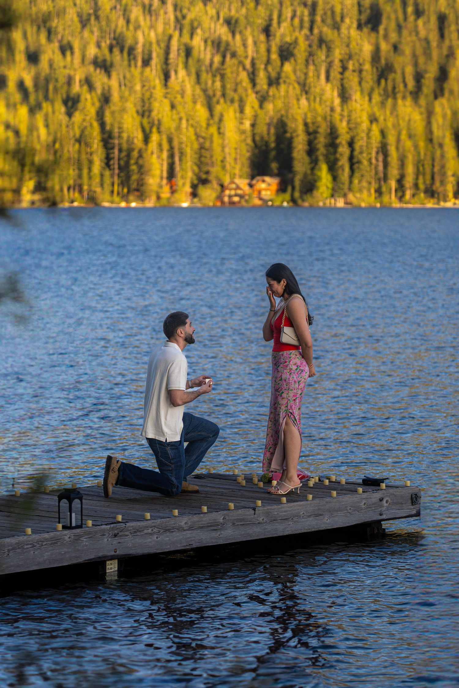 A romantic lakeside proposal on a wooden dock with lanterns and mountains in the background at sunset.