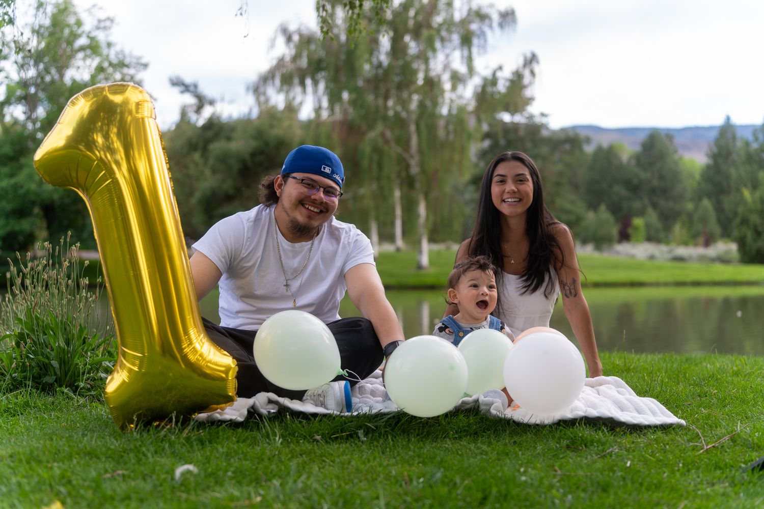 Family celebrates first birthday outdoors with gold number one balloon and white decorations by a lake.