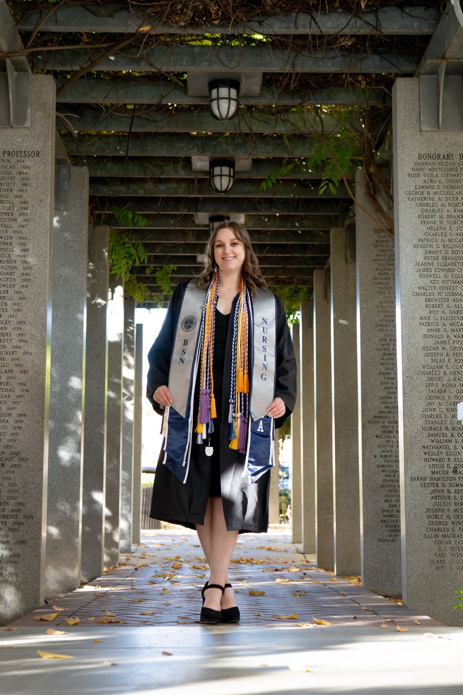 Graduate in black gown and stoles poses in sunlit columned walkway.