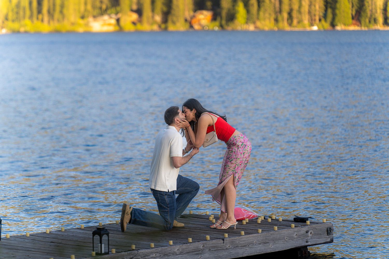A romantic proposal scene on a wooden dock by a lake with golden autumn trees in the background.