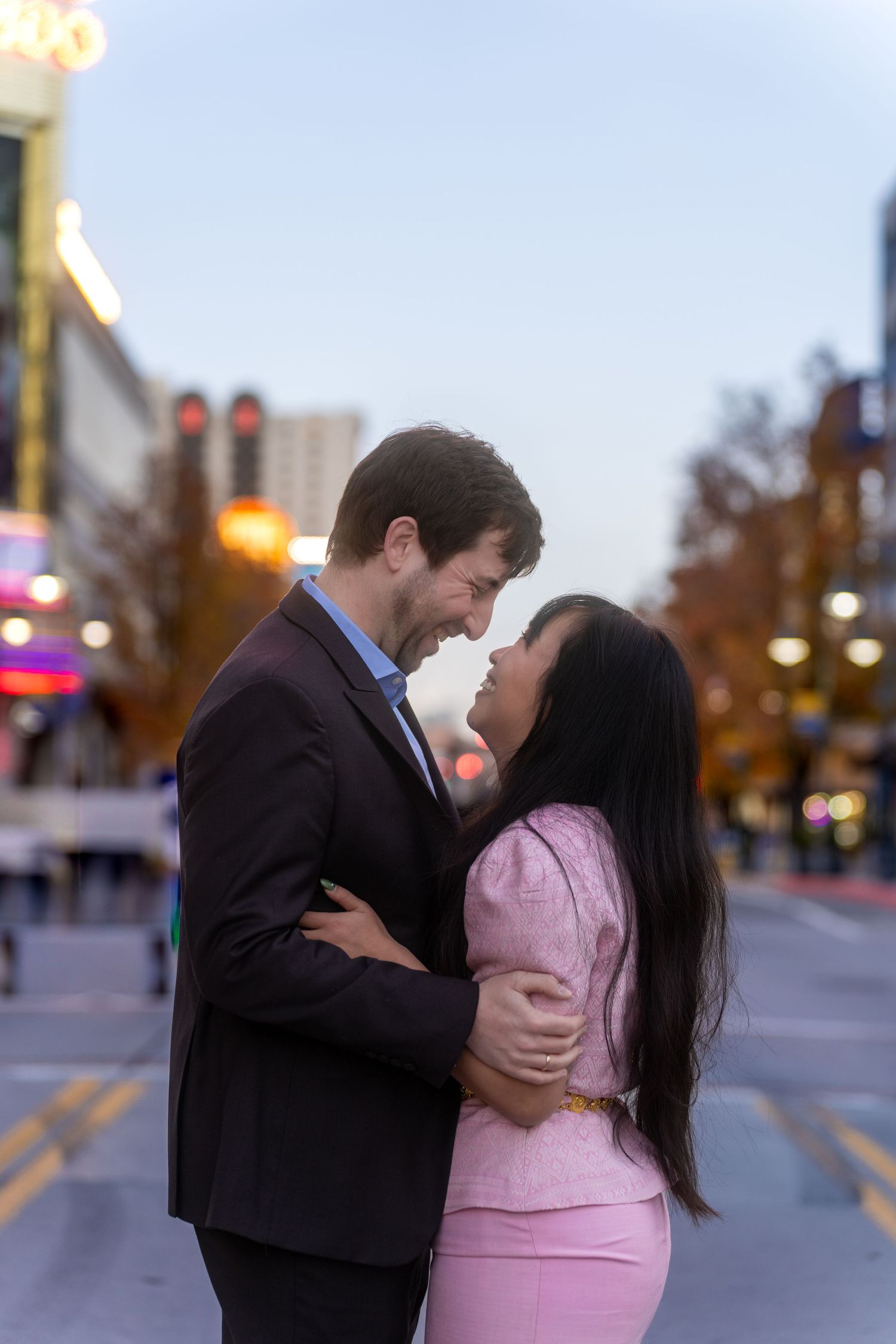 A romantic couple embraces in a city street at dusk, with the person in pink and black formal attire.