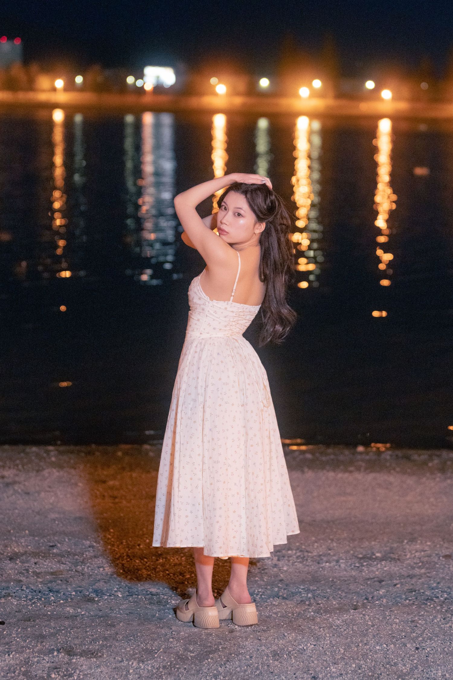A person in a white polka dot dress poses gracefully by the water at night with glowing lights reflecting on the surface.