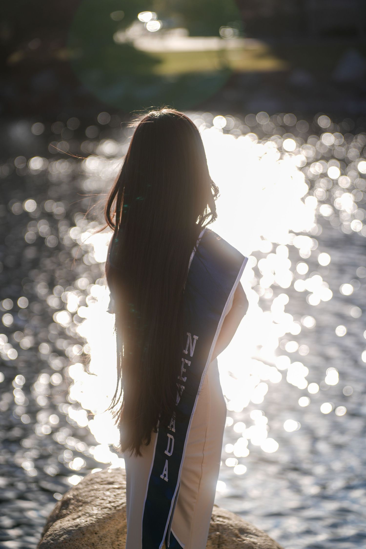 Silhouetted figure with graduation stole stands by sparkling water at sunset.