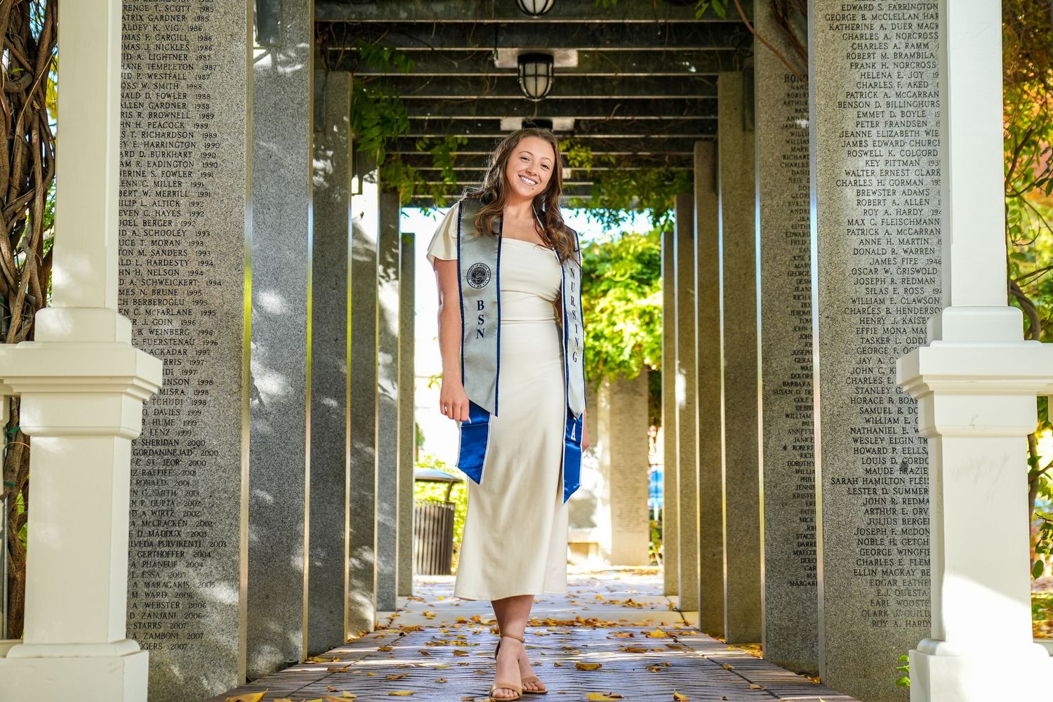 Graduate in white dress poses under architectural colonnade with hanging vines.