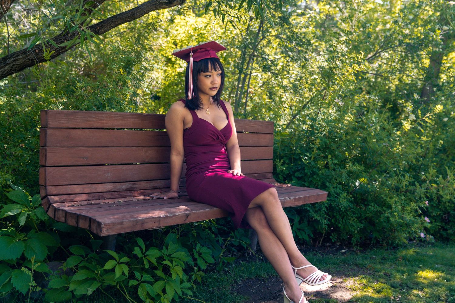 Graduate in burgundy dress sitting on wooden bench surrounded by lush greenery.
