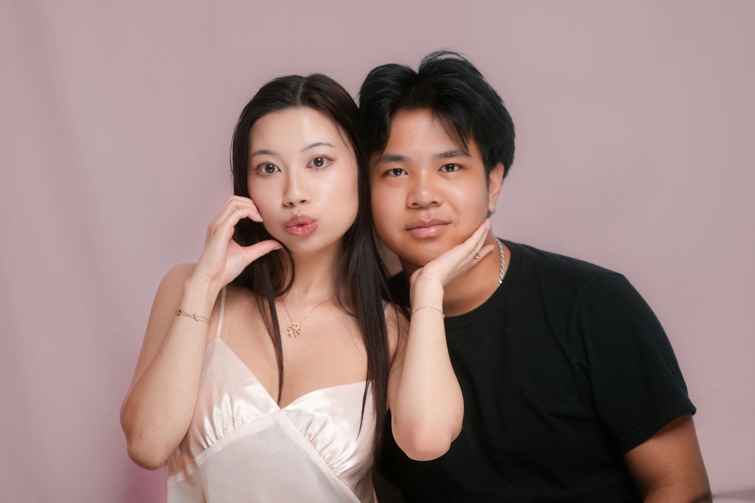 Two people in relaxed poses make playful expressions while taking a portrait photo against a pink backdrop.