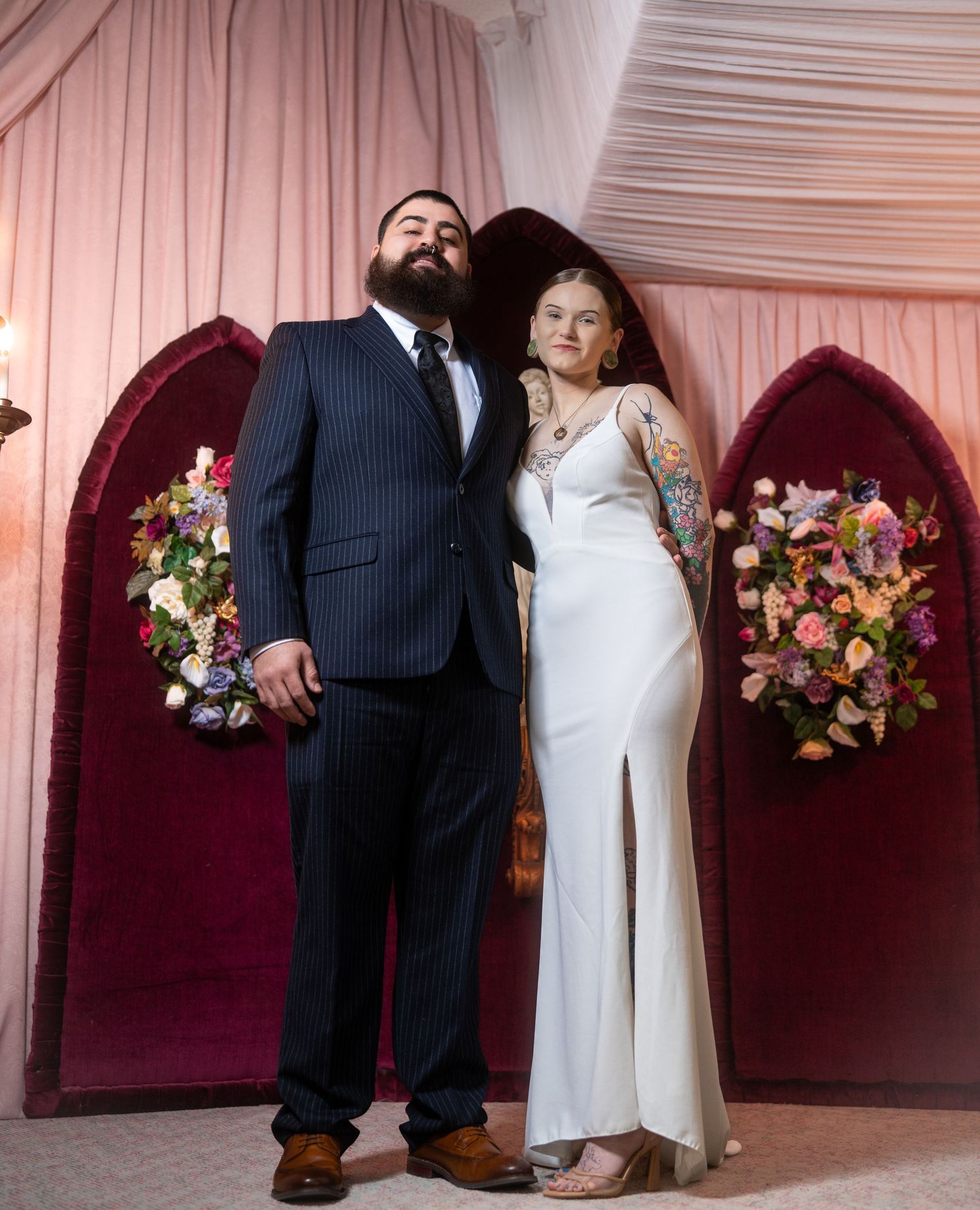 A couple in wedding attire poses in front of burgundy velvet panels decorated with floral arrangements.