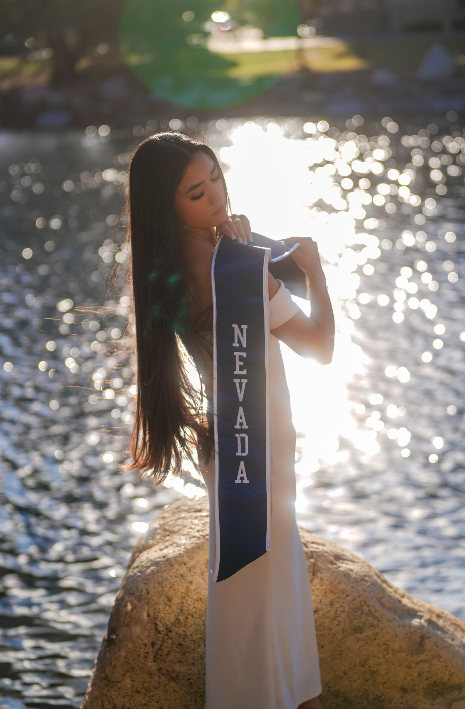 Graduate in white dress holds Nevada graduation stole by waterfront at golden hour.