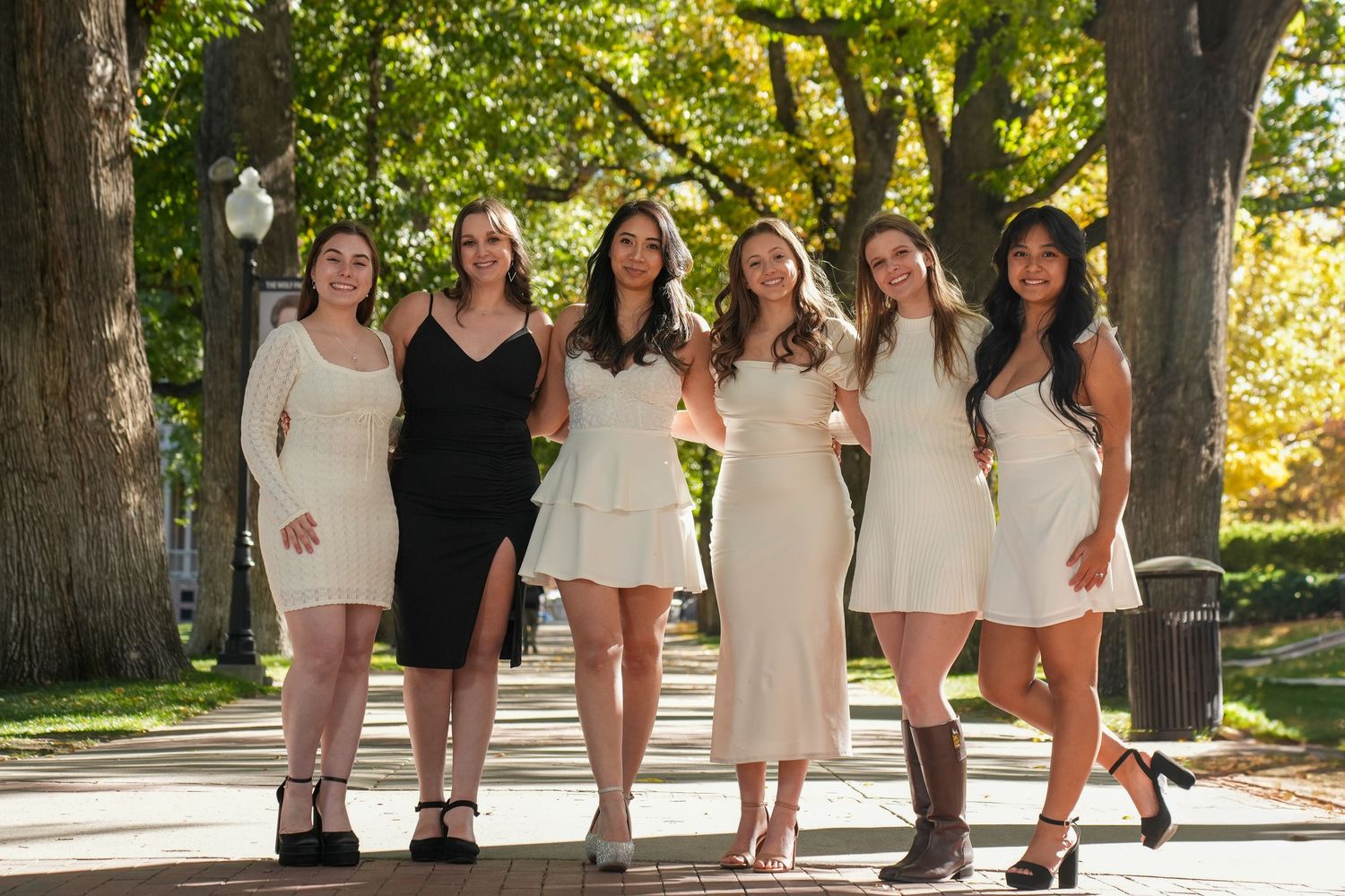 Group of women in white and black dresses posing together on tree-lined pathway.