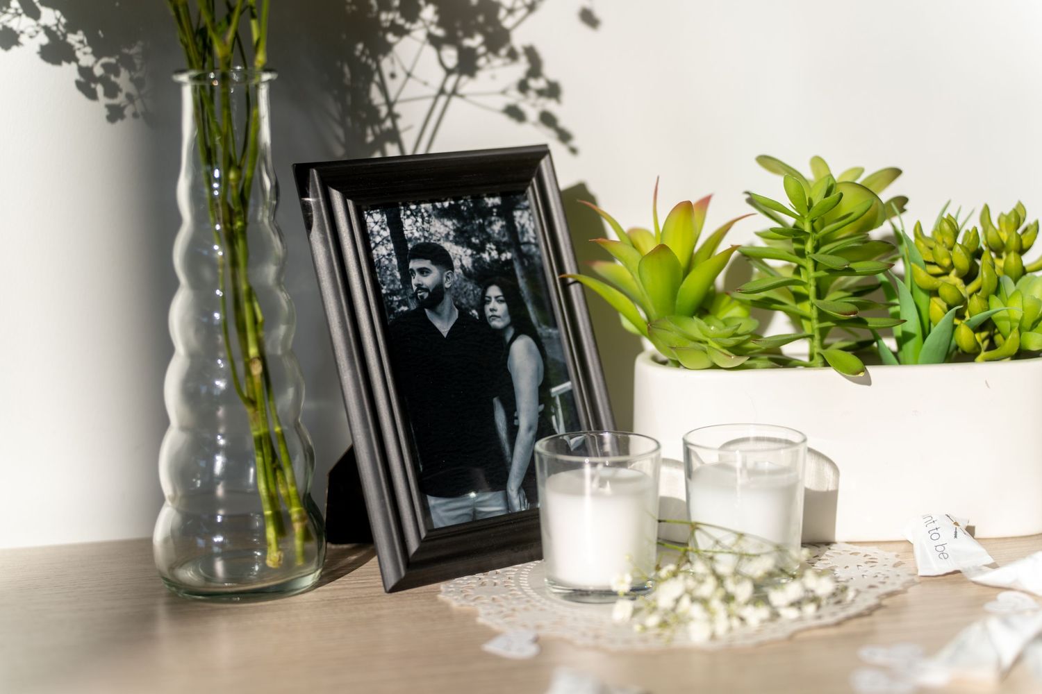 Decorative vase filled with fresh lily of the valley flowers sits on a white shelf with potted plants and framed artwork.