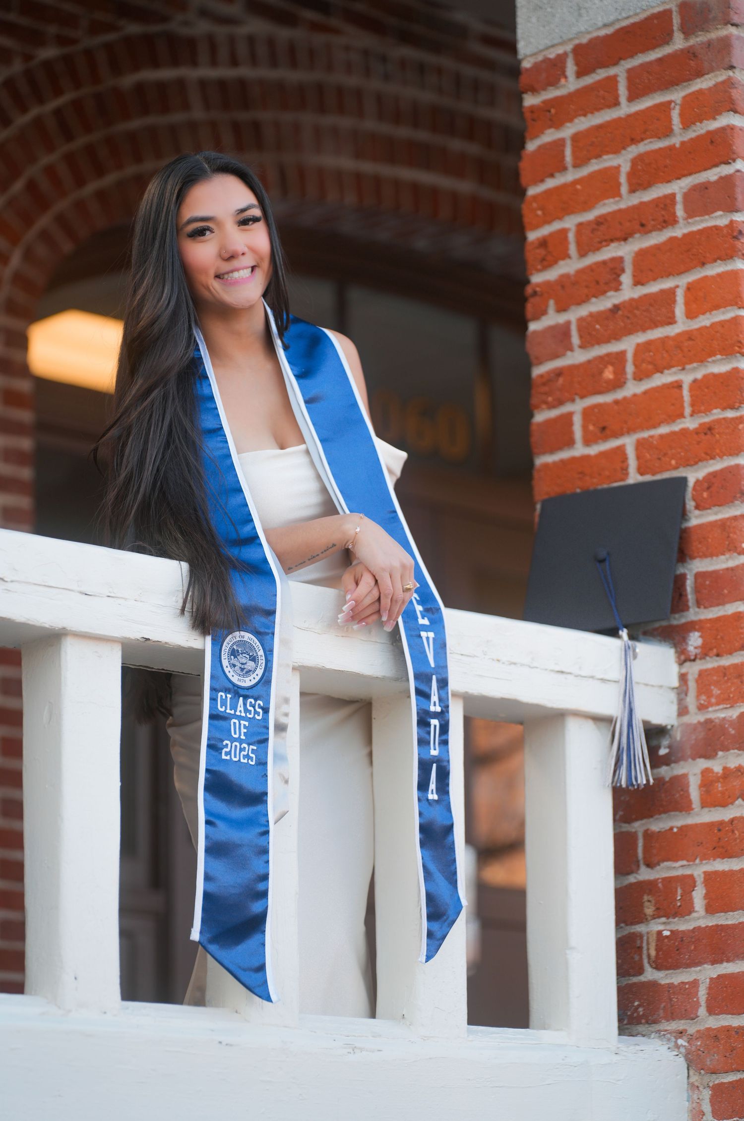 Smiling graduate with blue Nevada stole poses on white balcony with brick background.