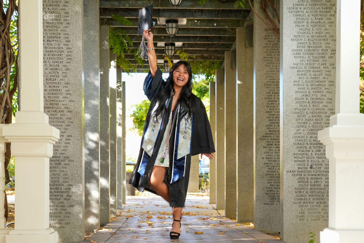 Graduate in cap and gown jumping joyfully under columned walkway.