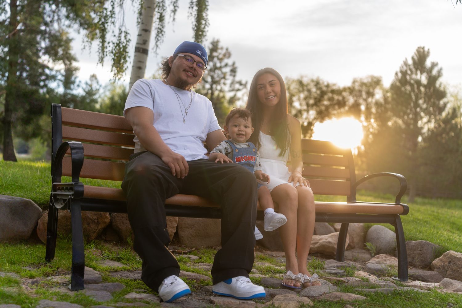 A family sits together on a wooden park bench at sunset with trees and golden sunlight in the background.