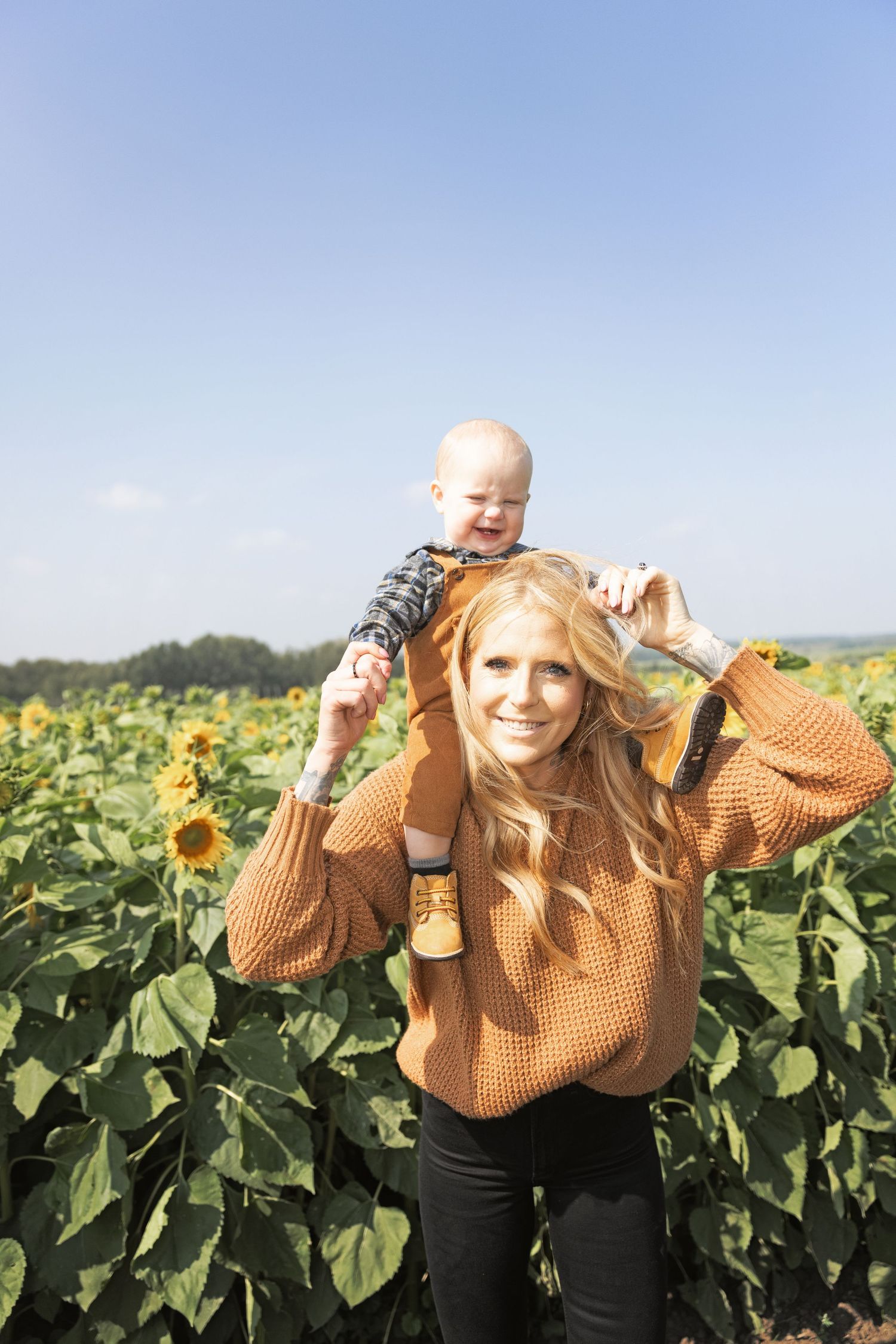 A woman stands in front of a field of sunflowers with a 1 year old child on her shoulders.