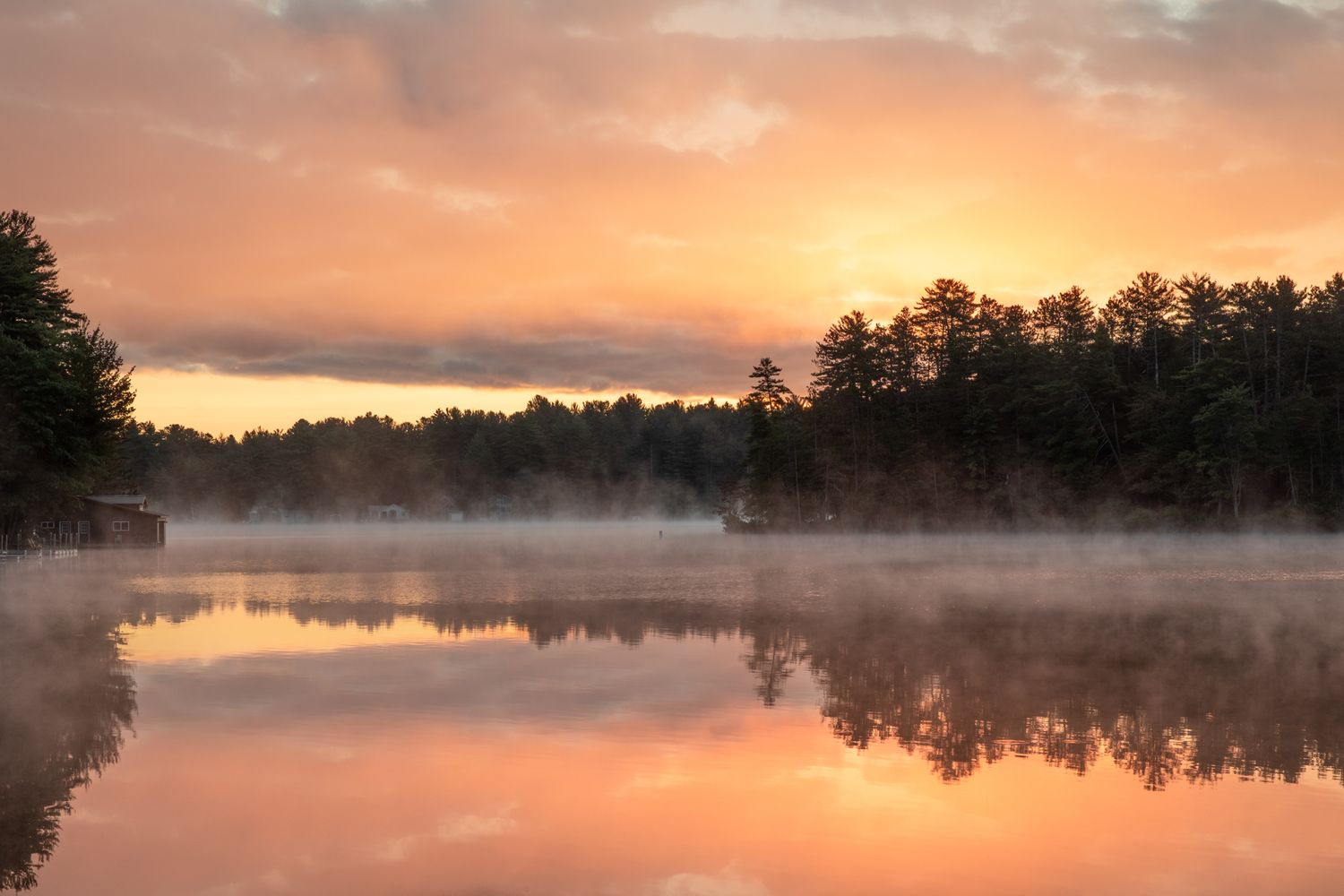 Misty lake at sunrise with orange sky reflected in perfectly still water surrounded by pine trees.