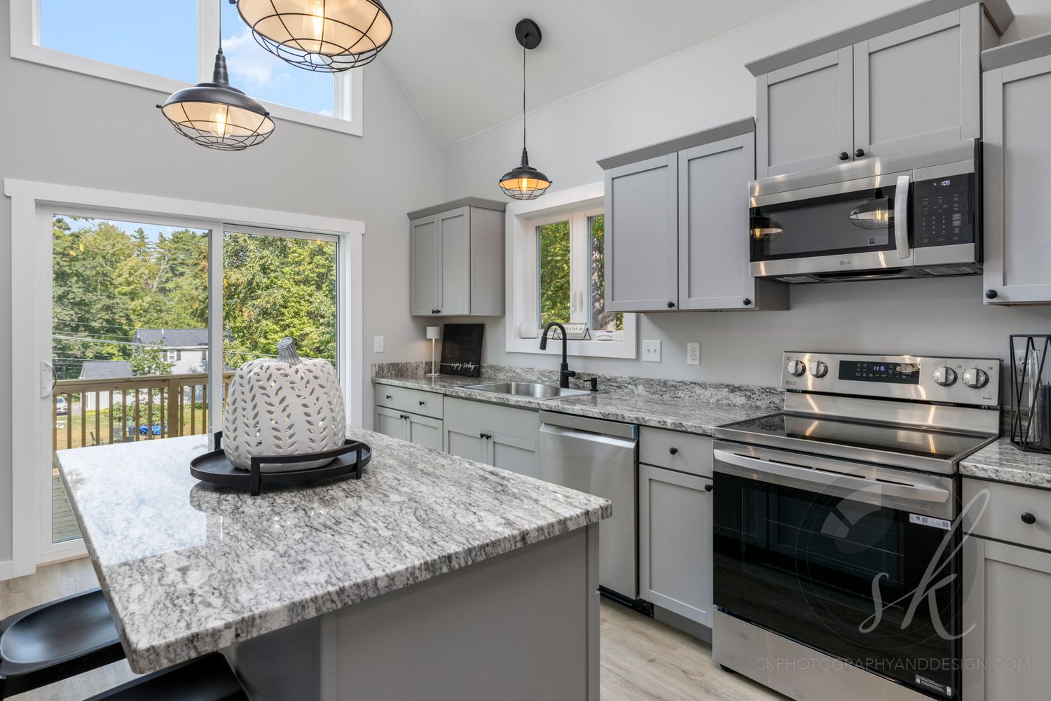 Modern kitchen with gray cabinets, granite island, stainless steel appliances, and large windows overlooking a deck.