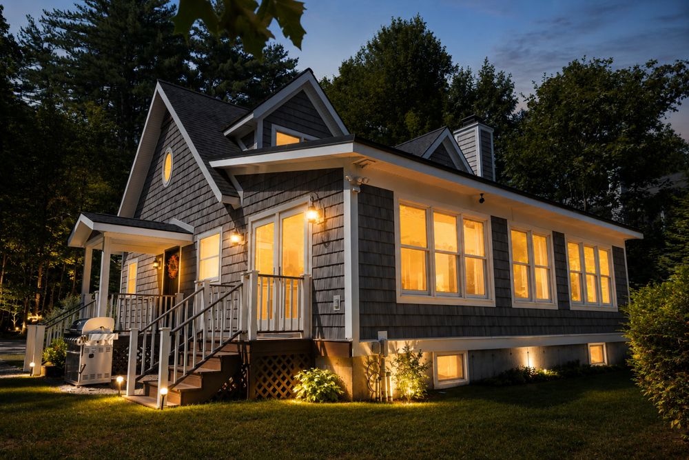 Twilight photo example of a charming gray shingle cottage in the Lake George area glowing at night.