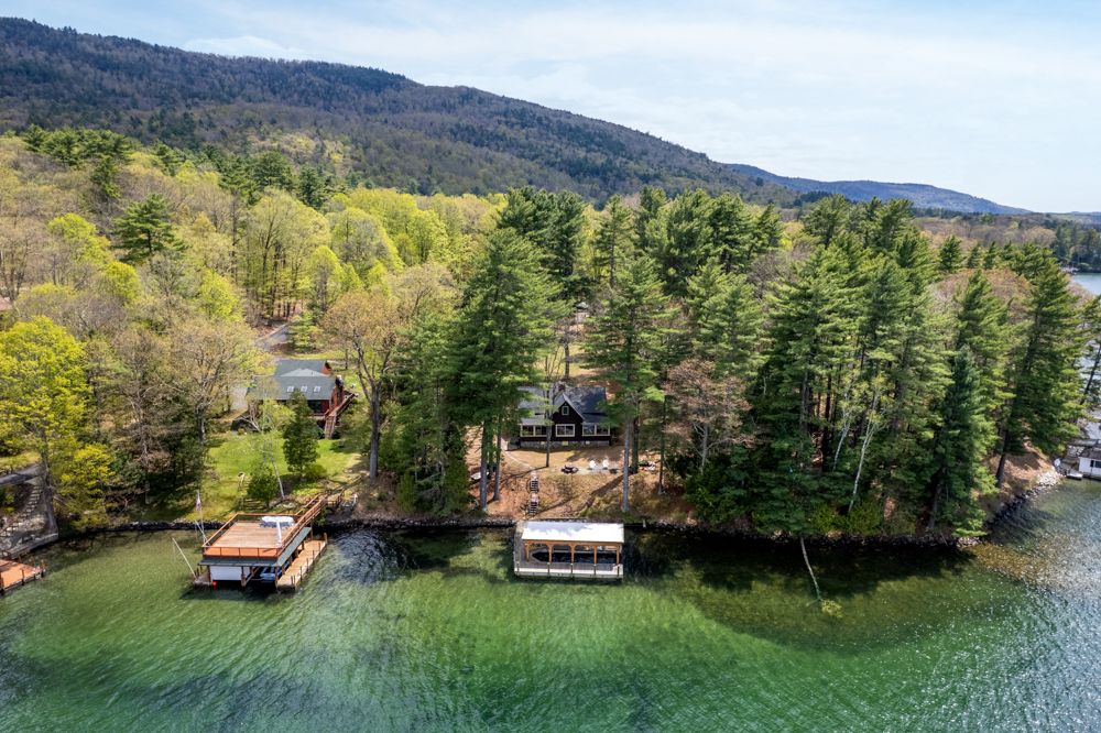 Aerial view of a lakeside cabin surrounded by tall pine trees with a private dock on calm green waters and mountain backdrop.