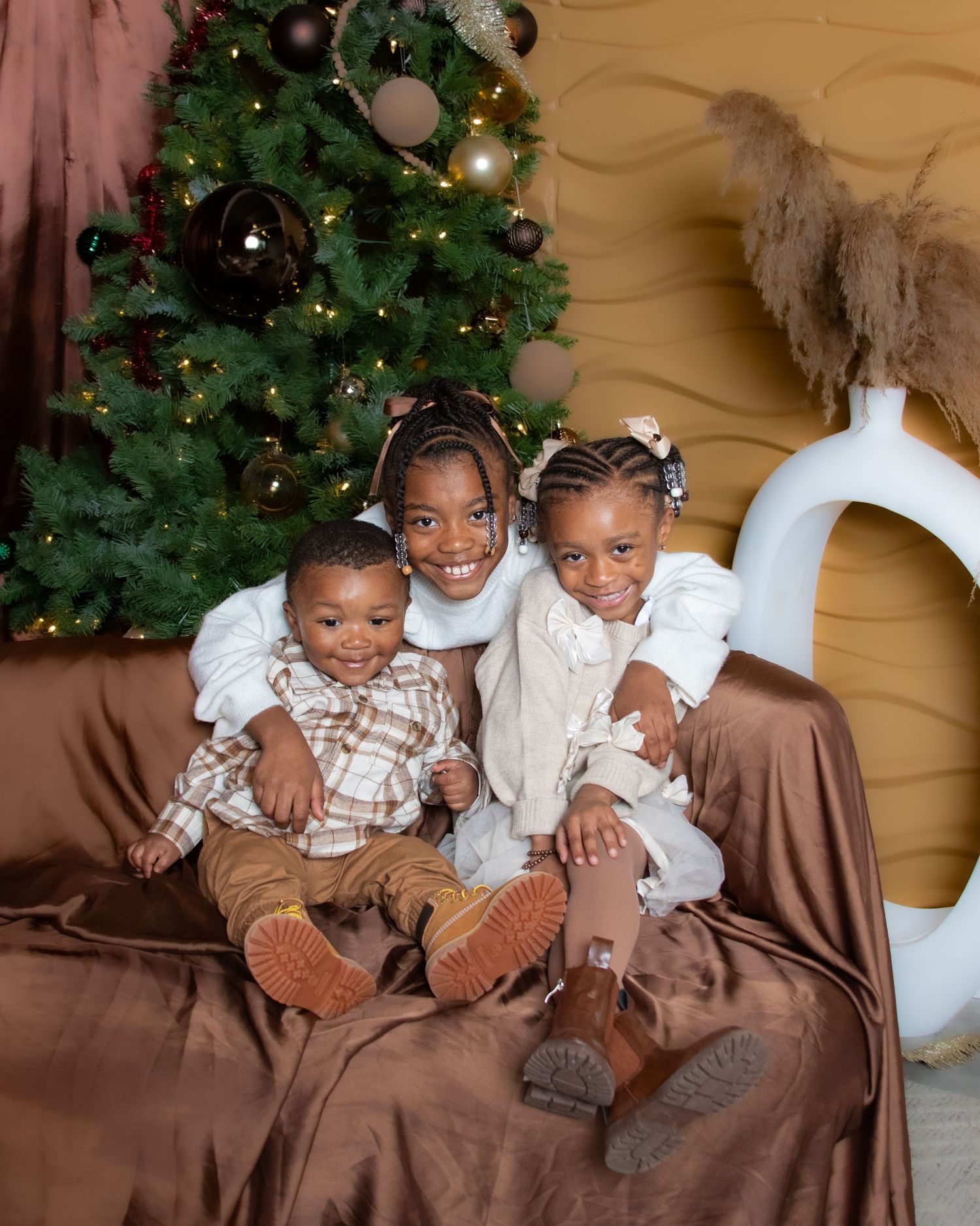 Three children sitting together on a brown couch with a decorated Christmas tree visible in the background.