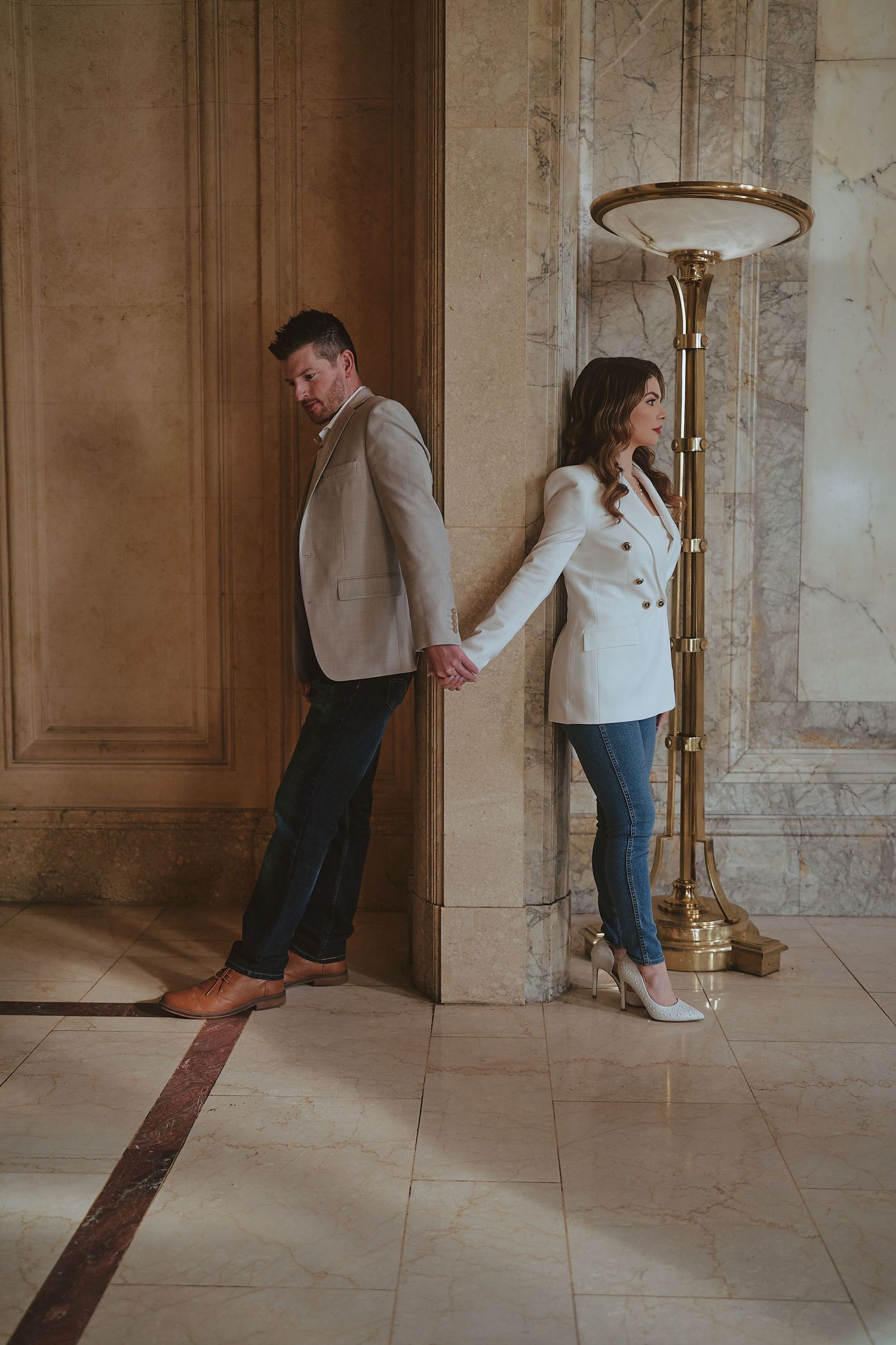 Couple holding hands around a marble column in an elegant building, dressed in blazers and jeans.