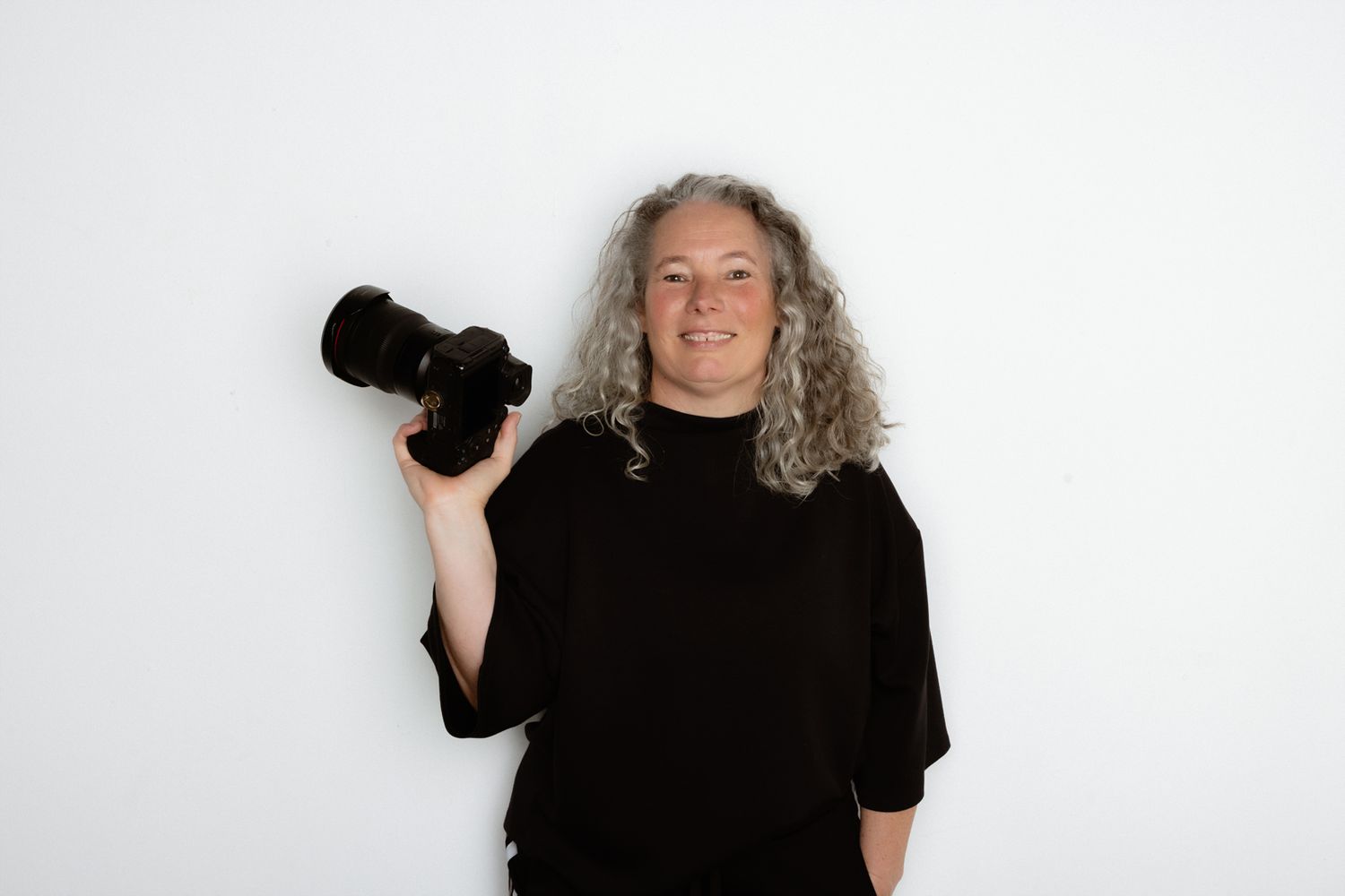 Smiling woman with curly gray hair holding a professional camera, wearing black top against white background.